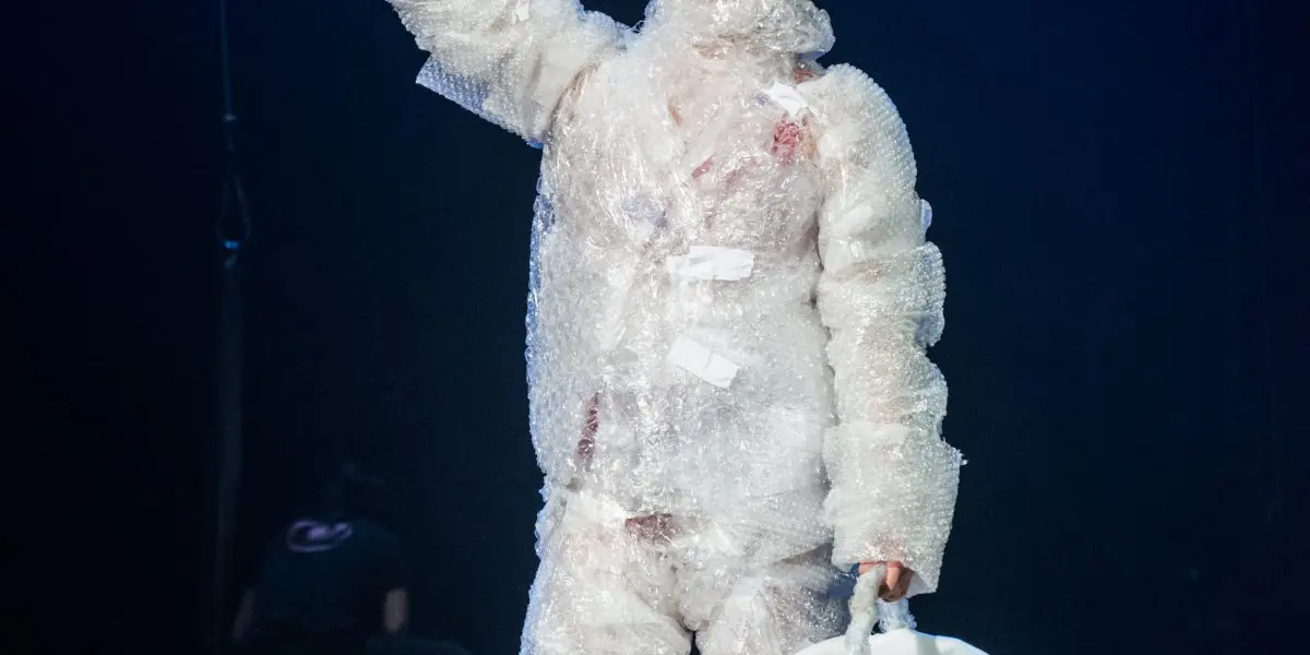 Une artiste en costume de papier bulle, peut-être Éline Guélat, tient un petit drapeau et un seau blanc sur une scène faiblement éclairée, tandis qu'une autre personne apparaît de façon floue à l'arrière-plan.