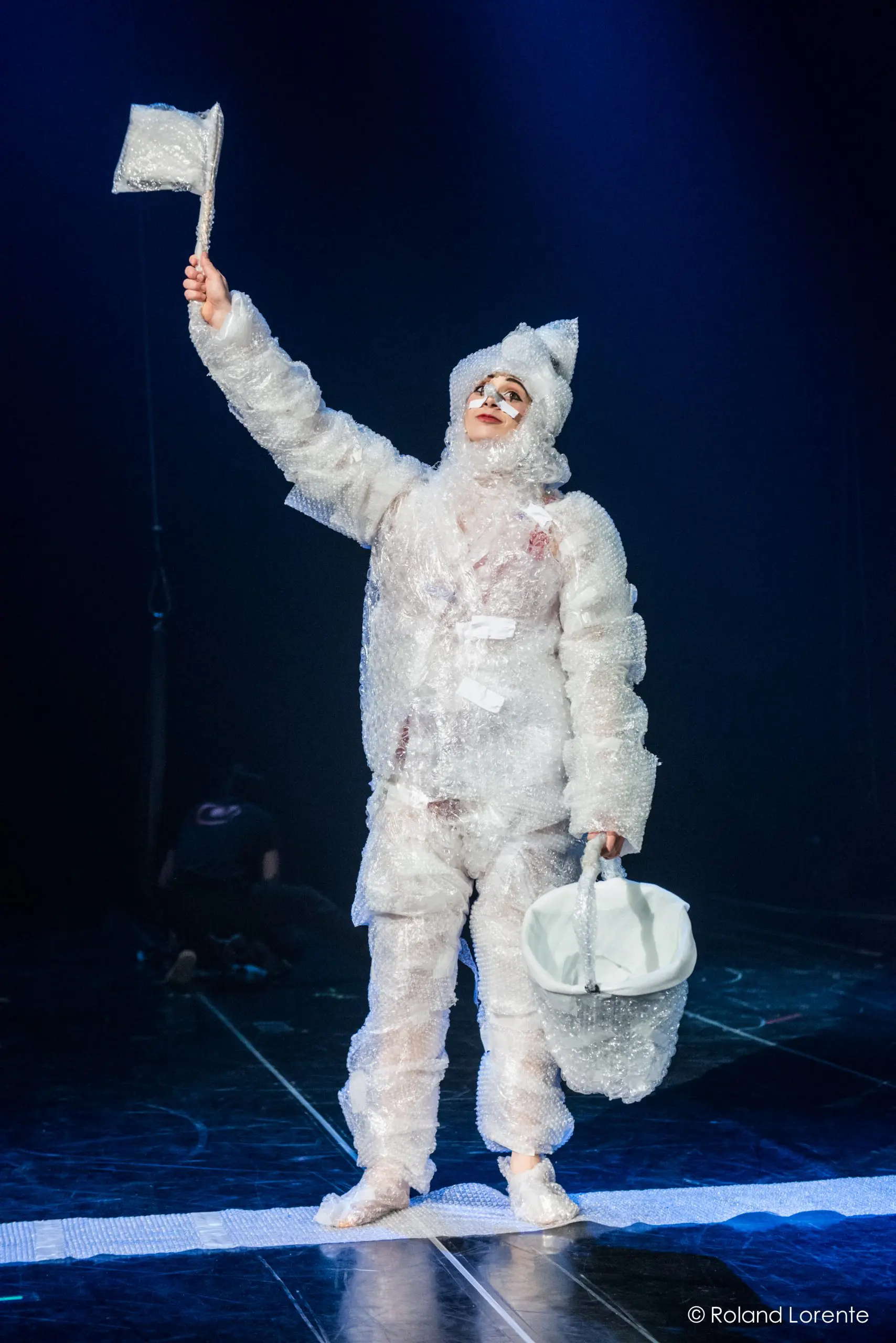 Une artiste en costume de papier bulle, peut-être Éline Guélat, tient un petit drapeau et un seau blanc sur une scène faiblement éclairée, tandis qu'une autre personne apparaît de façon floue à l'arrière-plan.