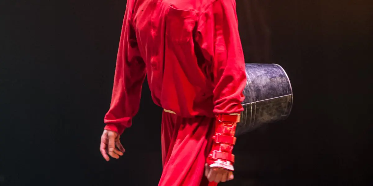 Éline Guélat, vêtue d'une tenue rouge vif, de chaussures assorties et de maquillage, se tient sur scène, souriante, en tenant un seau métallique derrière elle. Un éclairage dramatique la met en valeur sur un fond sombre.