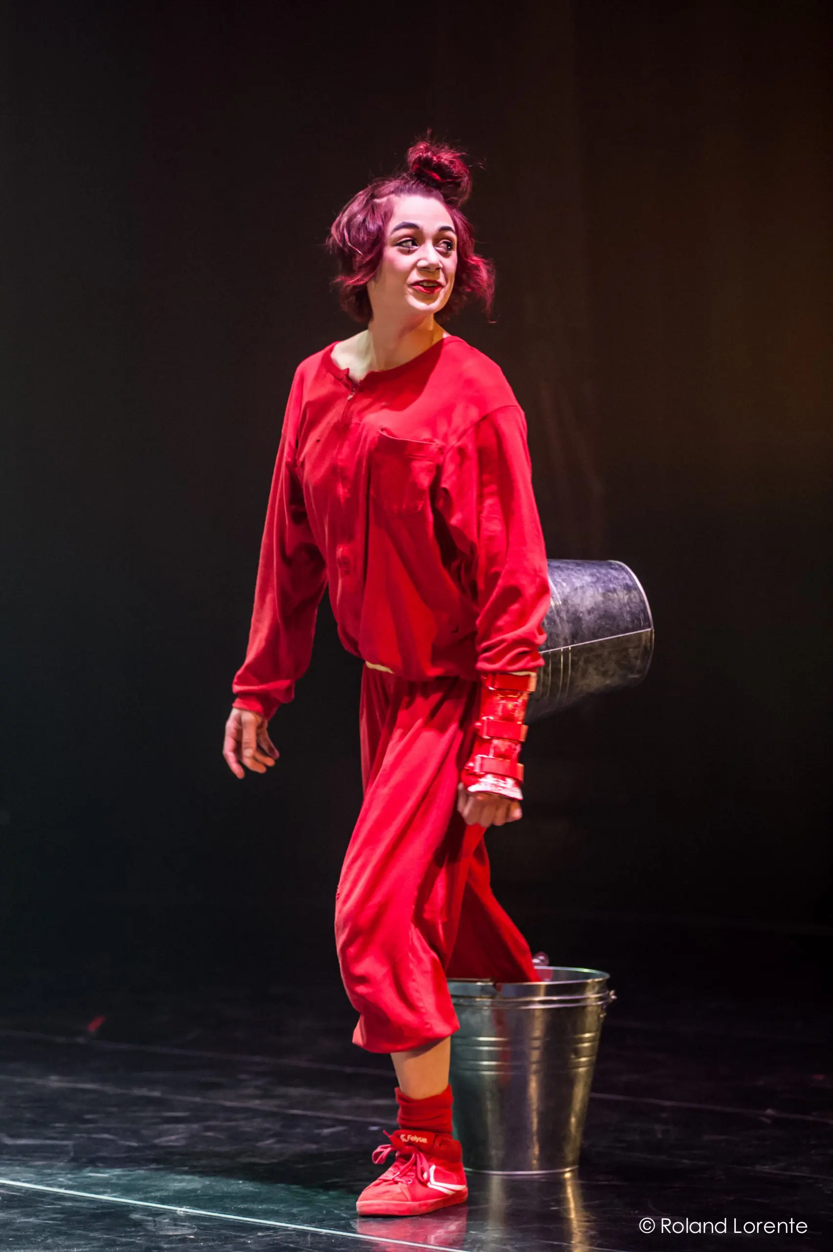 Éline Guélat, vêtue d'une tenue rouge vif, de chaussures assorties et de maquillage, se tient sur scène, souriante, en tenant un seau métallique derrière elle. Un éclairage dramatique la met en valeur sur un fond sombre.