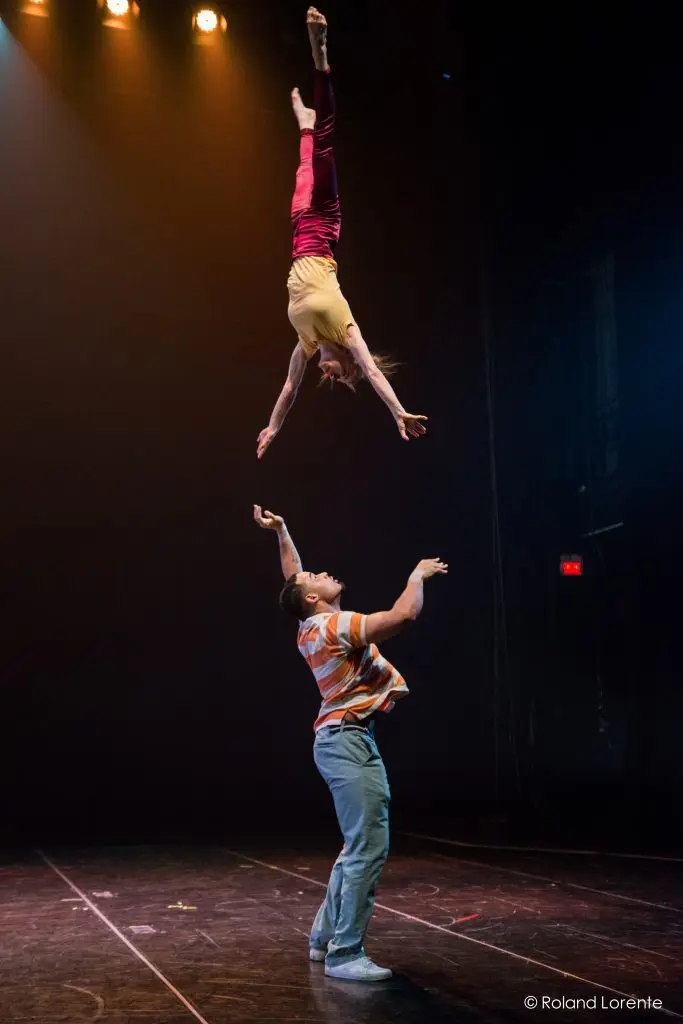 Un acrobate en chemise rayée et pantalon bleu, probablement Michael Patterson, fait tenir Joëlle Ziorjen en équilibre sur une main pendant une représentation sur scène, avec un éclairage dramatique au-dessus de la tête.