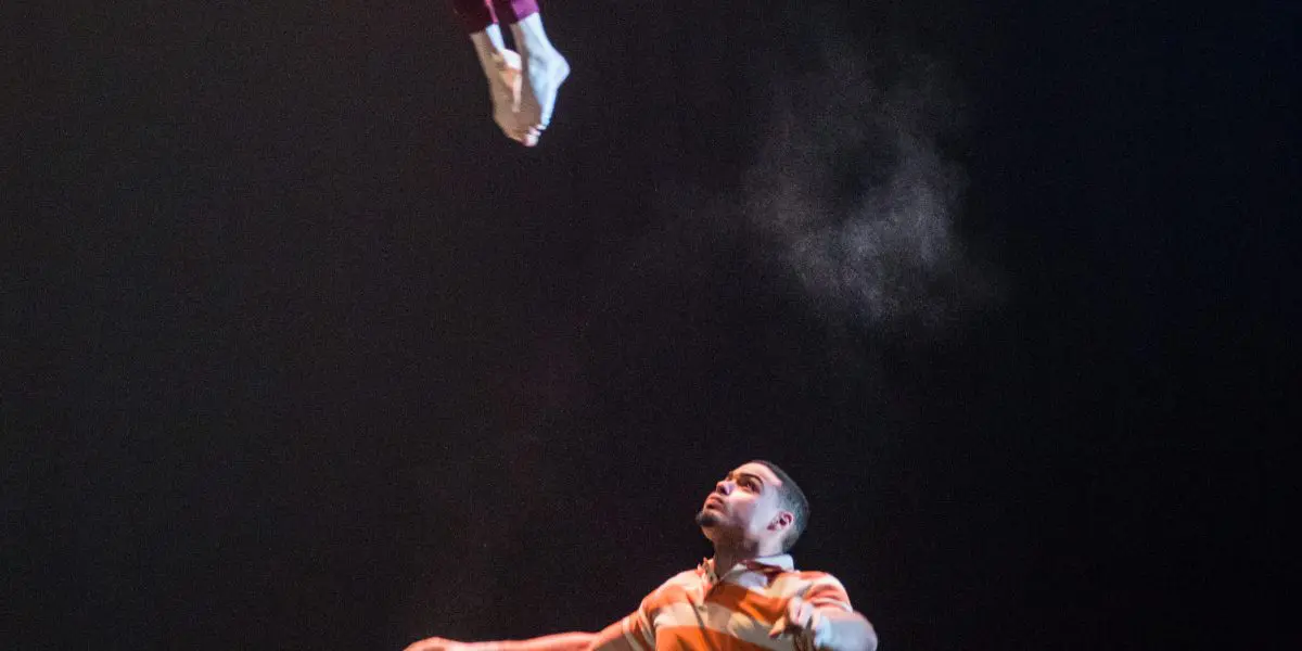 Joëlle Ziorjen et Michael Patterson se produisent sous les lumières de la scène : Joëlle est capturée en plein vol, les jambes pliées dans un saut gracieux au-dessus de Michael, qui se tient en bas dans une chemise rayée, regardant vers le haut sur le fond sombre.