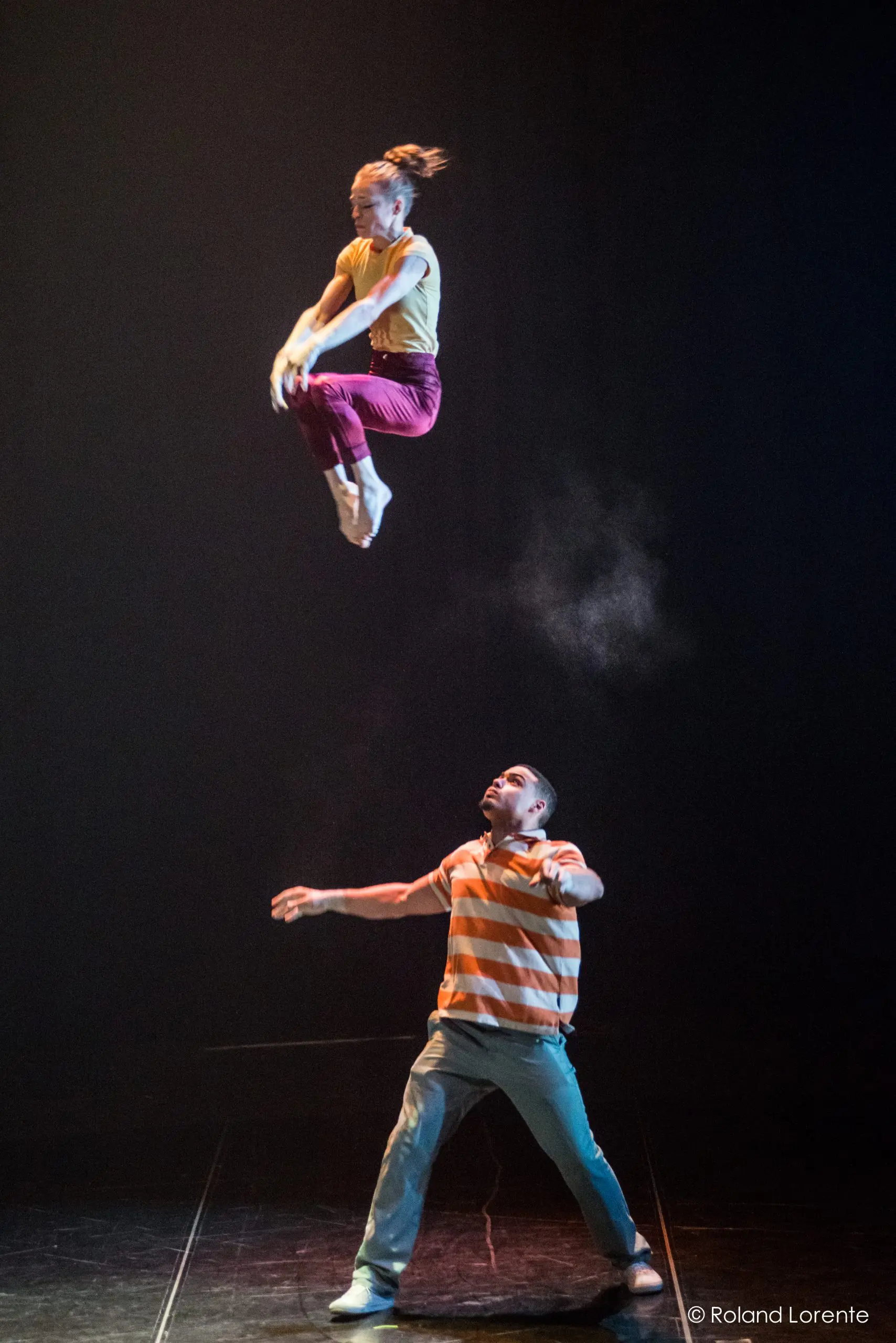 Joëlle Ziorjen et Michael Patterson se produisent sous les lumières de la scène : Joëlle est capturée en plein vol, les jambes pliées dans un saut gracieux au-dessus de Michael, qui se tient en bas dans une chemise rayée, regardant vers le haut sur le fond sombre.