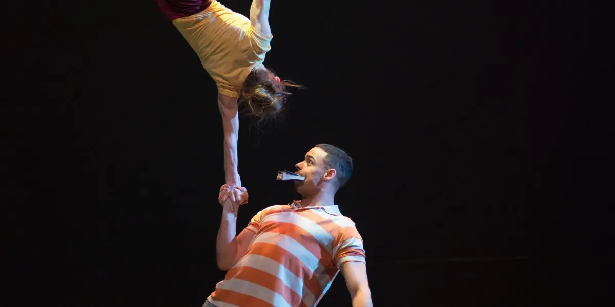 Joëlle Ziorjen et Michael Patterson éblouissent alors qu'un homme en chemise rayée fait tenir une femme à l'envers sur une main lors d'une performance acrobatique sur une scène sombre.