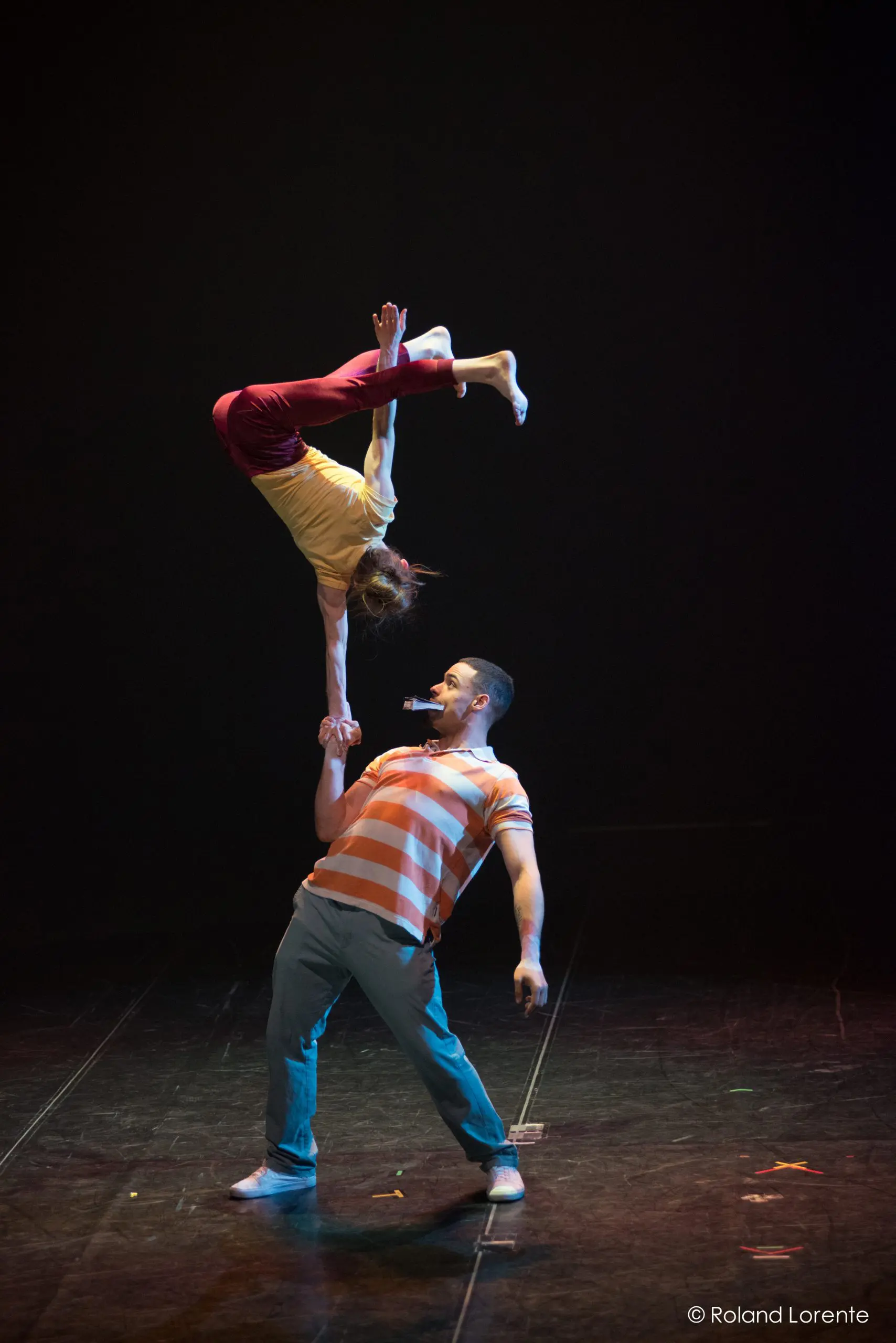 Joëlle Ziorjen et Michael Patterson éblouissent alors qu'un homme en chemise rayée fait tenir une femme à l'envers sur une main lors d'une performance acrobatique sur une scène sombre.