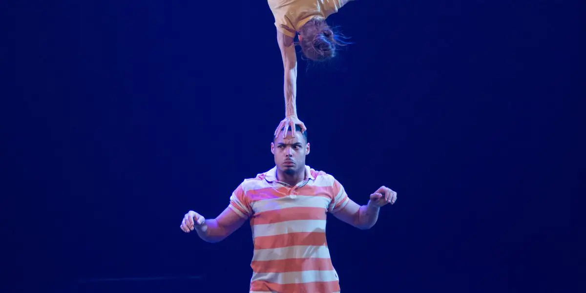 Joëlle Ziorjen et Michael Patterson éblouissent lorsqu'un acrobate masculin se tient en équilibre sur la scène tandis que sa partenaire effectue un équilibre sur une main sur sa tête, les jambes étendues en fente, le tout sous un éclairage bleu dramatique.