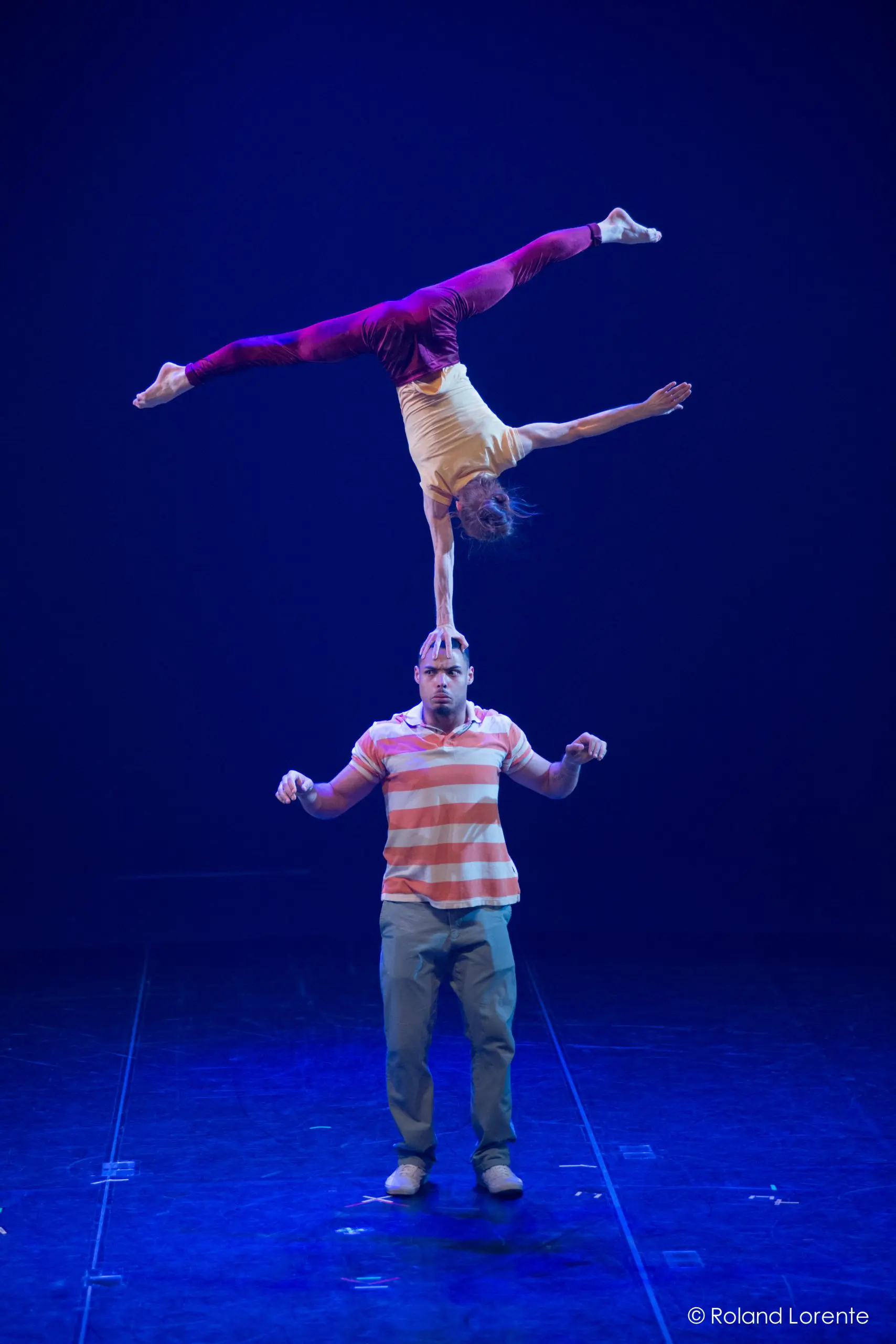 Joëlle Ziorjen et Michael Patterson éblouissent lorsqu'un acrobate masculin se tient en équilibre sur la scène tandis que sa partenaire effectue un équilibre sur une main sur sa tête, les jambes étendues en fente, le tout sous un éclairage bleu dramatique.