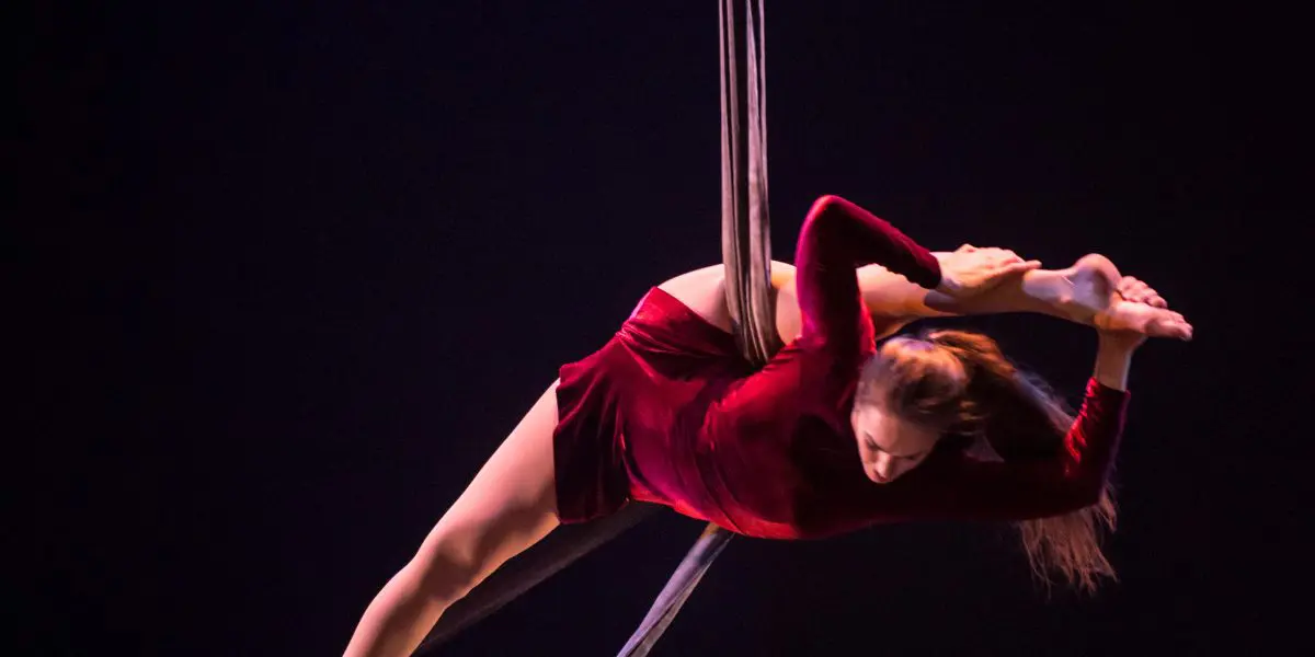 L'artiste aérienne Catherine Beaudet, vêtue d'une robe rouge, se tient gracieusement en équilibre sur un trapèze suspendu, en arquant le dos et en tenant son pied derrière la tête sur une scène sombre.