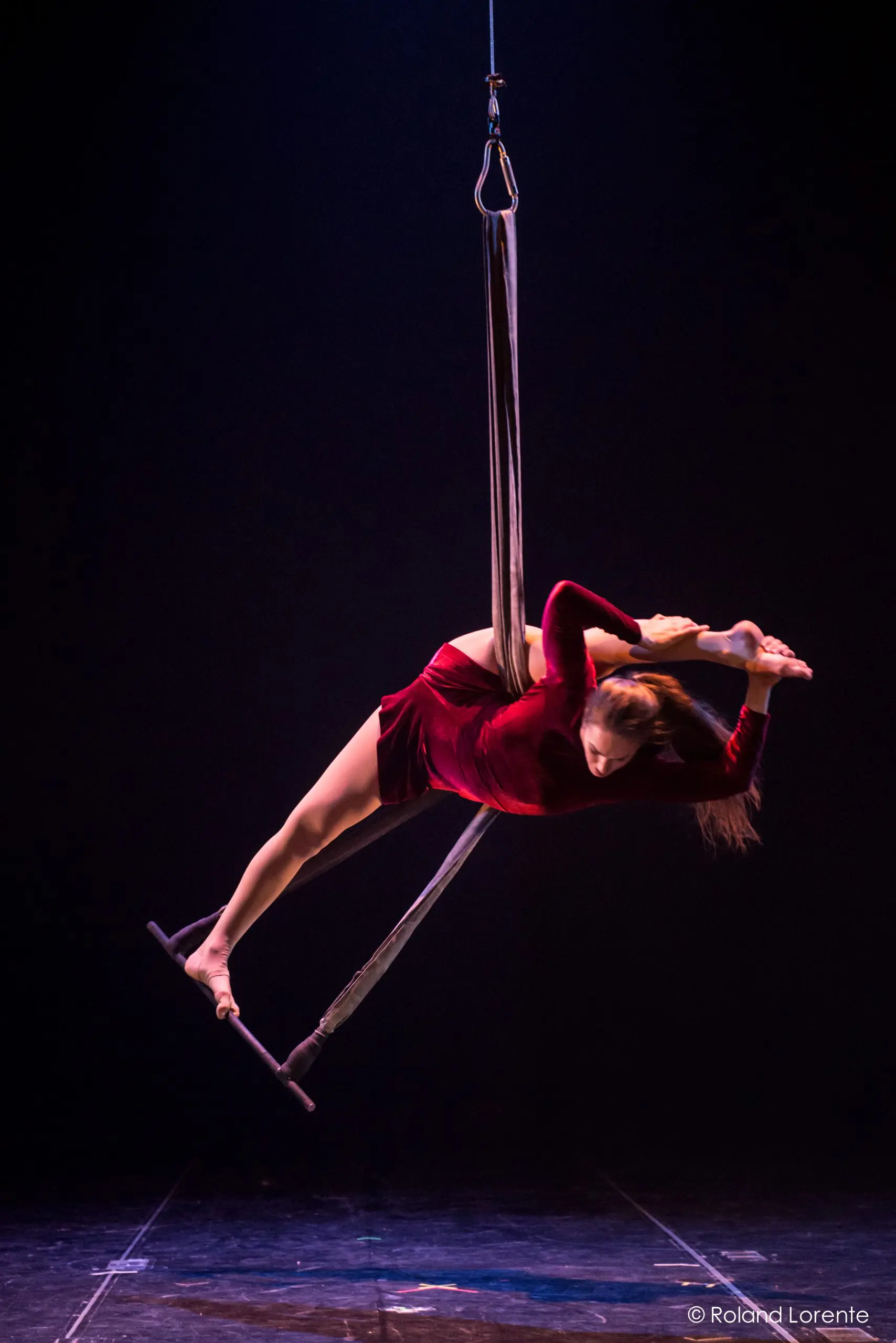 L'artiste aérienne Catherine Beaudet, vêtue d'une robe rouge, se tient gracieusement en équilibre sur un trapèze suspendu, en arquant le dos et en tenant son pied derrière la tête sur une scène sombre.