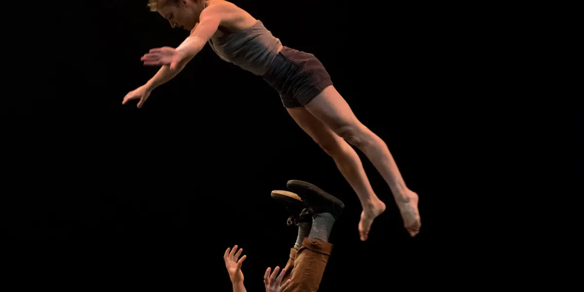 Andrei Anissimov et Emma Rogers se produisent alors qu'une danseuse s'envole, bras tendus, au-dessus d'un danseur allongé sur le dos, la soulevant avec ses pieds et ses mains lors d'un spectacle de danse contemporaine sur une scène faiblement éclairée.