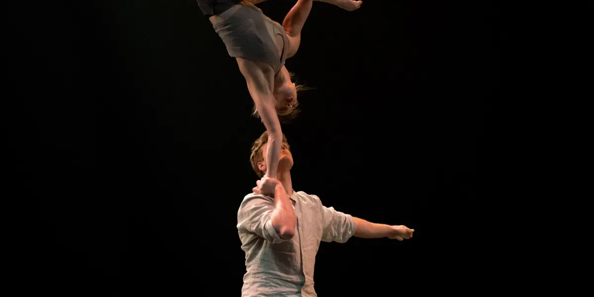Andrei Anissimov se tient sur scène et tient Emma Rogers en équilibre au-dessus de sa tête avec un bras. Elle est à l'envers, les jambes étendues et les bras tendus, sur un fond sombre.
