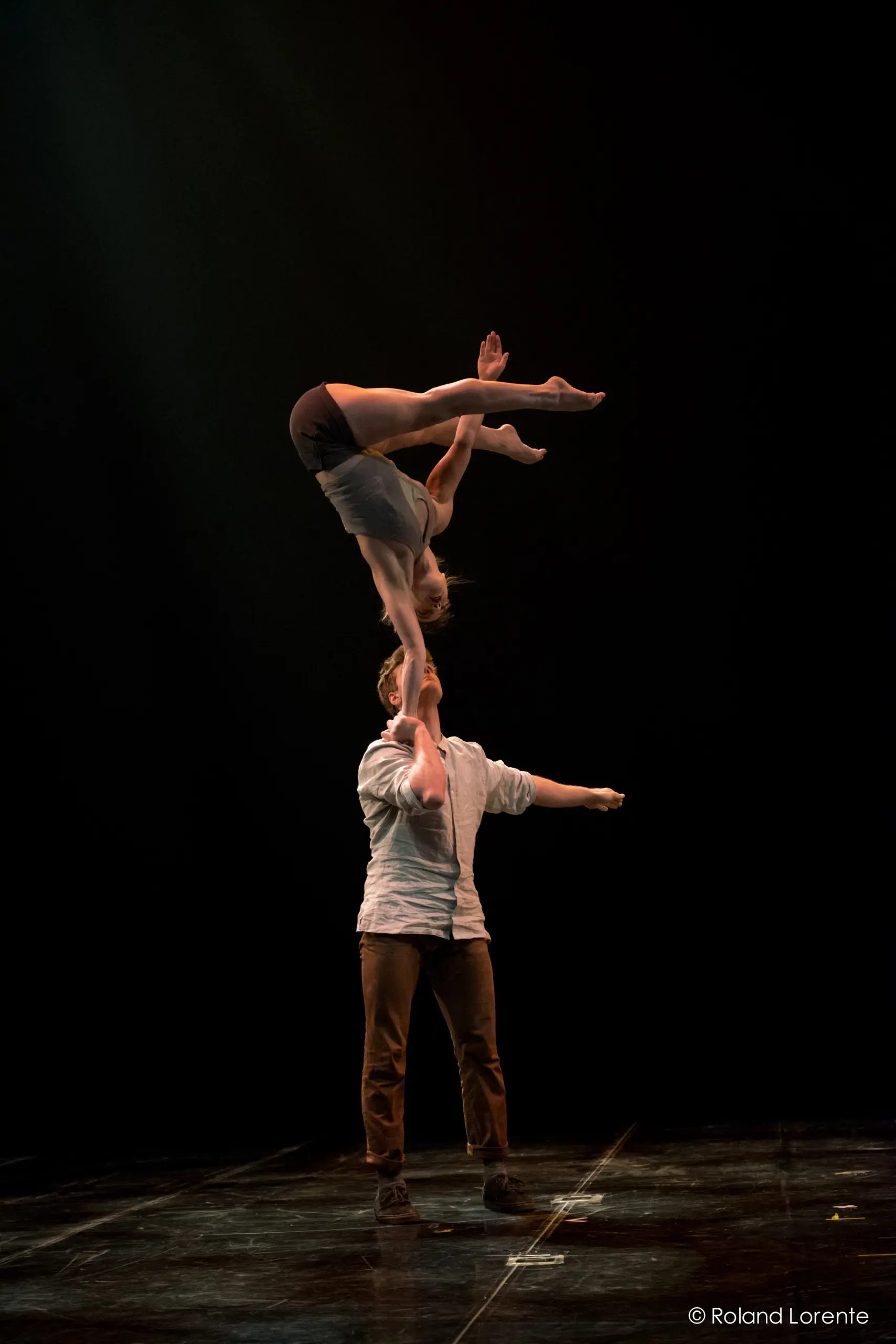 Andrei Anissimov se tient sur scène et tient Emma Rogers en équilibre au-dessus de sa tête avec un bras. Elle est à l'envers, les jambes étendues et les bras tendus, sur un fond sombre.