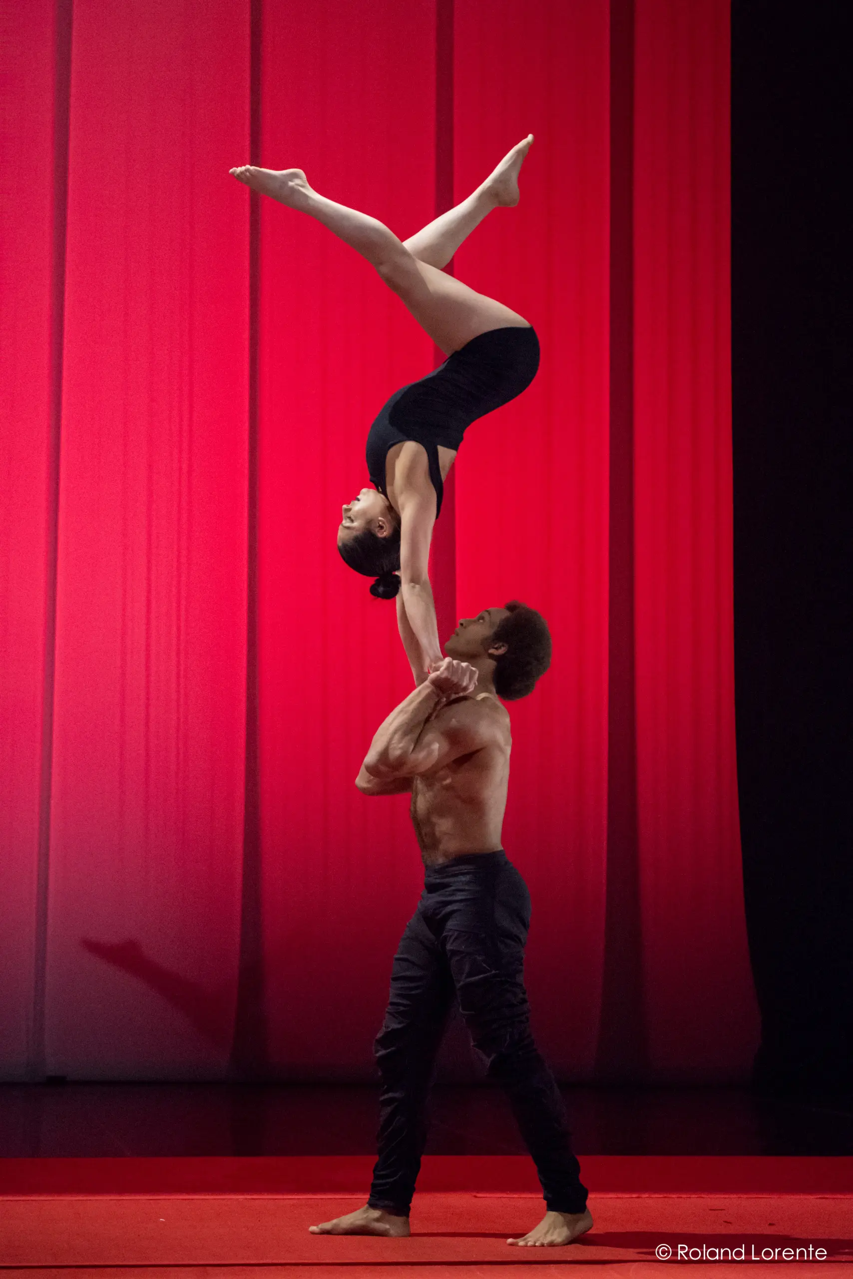 Marlon Archer fait tenir Jenay Espinosa en équilibre sur une main, la tête en bas, sur un fond de rideau rouge. Elle étend ses jambes en deux, tandis qu'il se tient fermement sur la scène rouge.