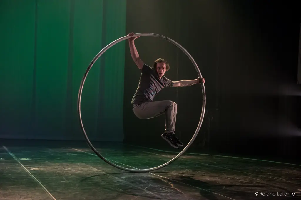 Cory Marsh se tient gracieusement en équilibre à l'intérieur d'une grande roue Cyr sur une scène faiblement éclairée, avec un éclairage vert qui projette des ombres dramatiques en arrière-plan.