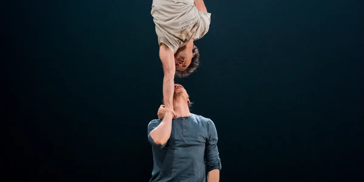 Deux interprètes, Julius Bitterling et César Mispelon, exécutent un mouvement acrobatique : l'un se tient debout, l'autre en équilibre sur une main, sous une scène sombre éclairée par un seul projecteur.