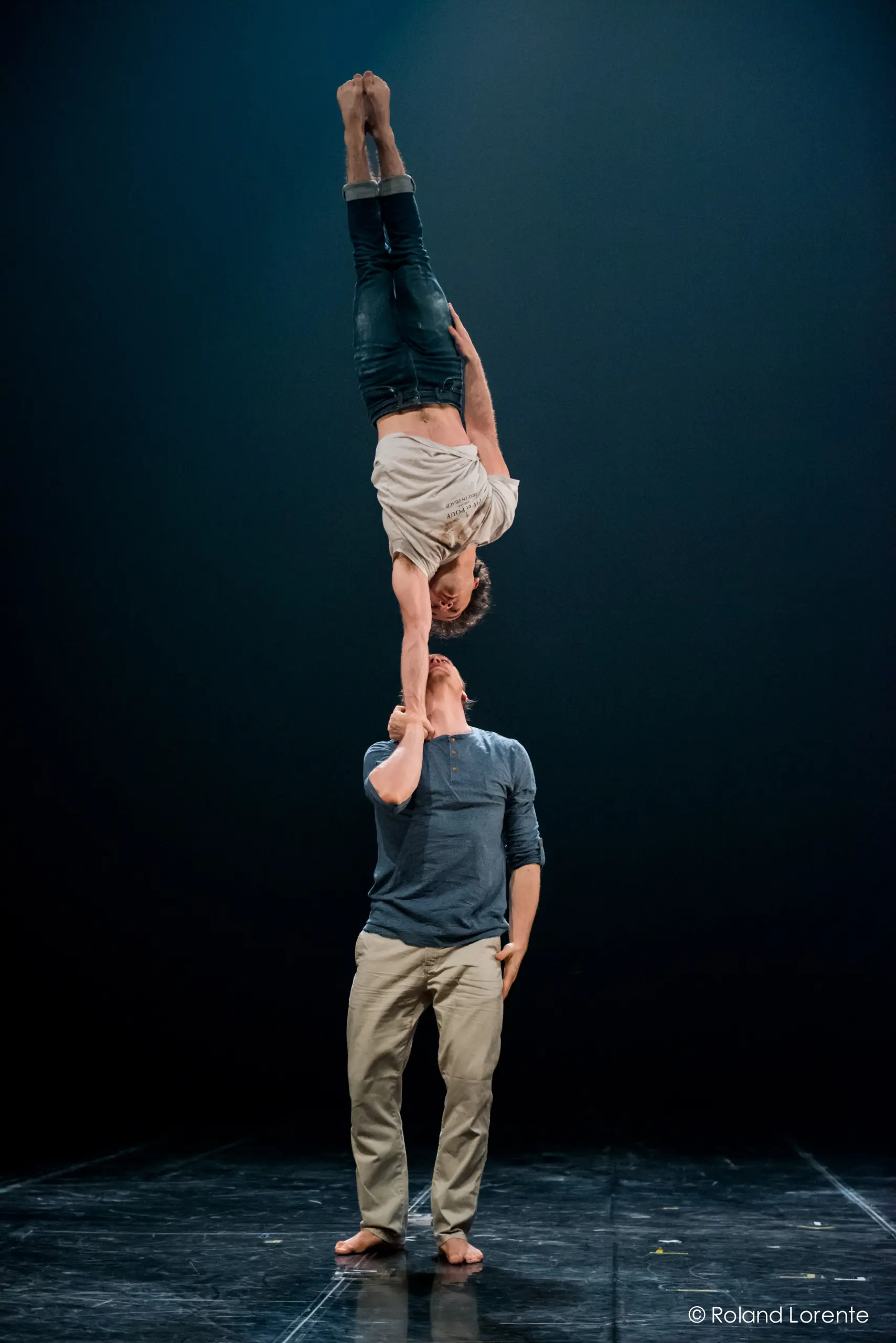 Deux interprètes, Julius Bitterling et César Mispelon, exécutent un mouvement acrobatique : l'un se tient debout, l'autre en équilibre sur une main, sous une scène sombre éclairée par un seul projecteur.
