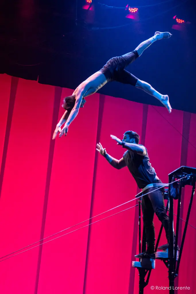 Un acrobate torse nu, Louis Joyal, est en plein vol, plongeant vers Samuel Renaud qui se tient sur une plate-forme, tous deux placés sur un fond de scène rose et violet vibrant.