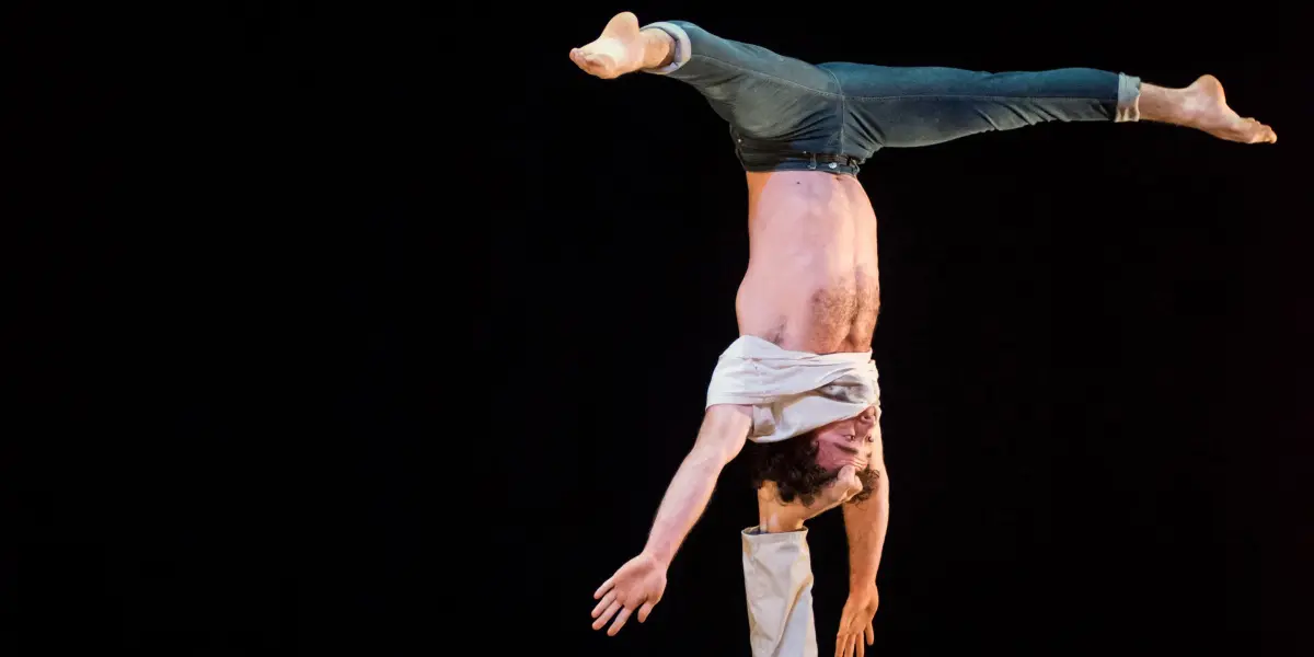 Deux acrobates masculins, Julius Bitterling et César Mispelon, se produisent sur scène ; l'un est allongé sur le dos et tient l'autre en équilibre sur un bras au-dessus de lui, la tête en bas. L'arrière-plan sombre met en valeur leur force et leur équilibre.