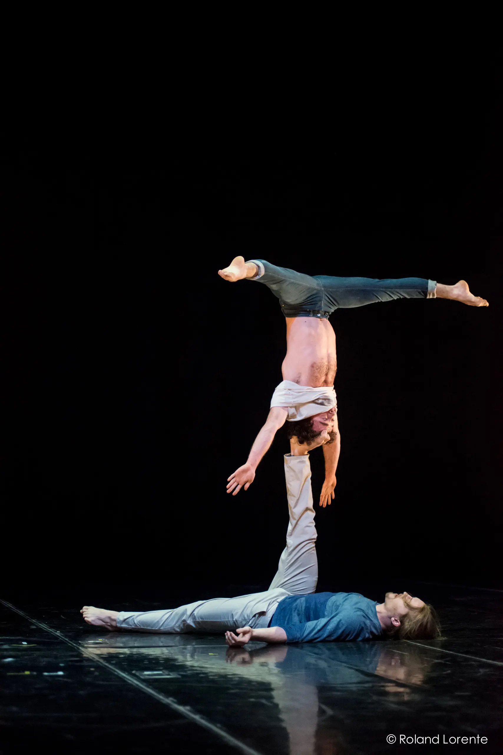 Deux acrobates masculins, Julius Bitterling et César Mispelon, se produisent sur scène ; l'un est allongé sur le dos et tient l'autre en équilibre sur un bras au-dessus de lui, la tête en bas. L'arrière-plan sombre met en valeur leur force et leur équilibre.