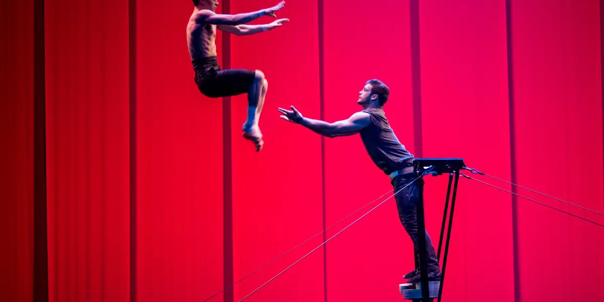 Deux acrobates, Louis Joyal et Samuel Renaud, réalisent une acrobatie : l'un saute dans les airs vers l'autre, qui se tient sur une plate-forme surélevée, les bras tendus, sur un fond rose-rouge vibrant.