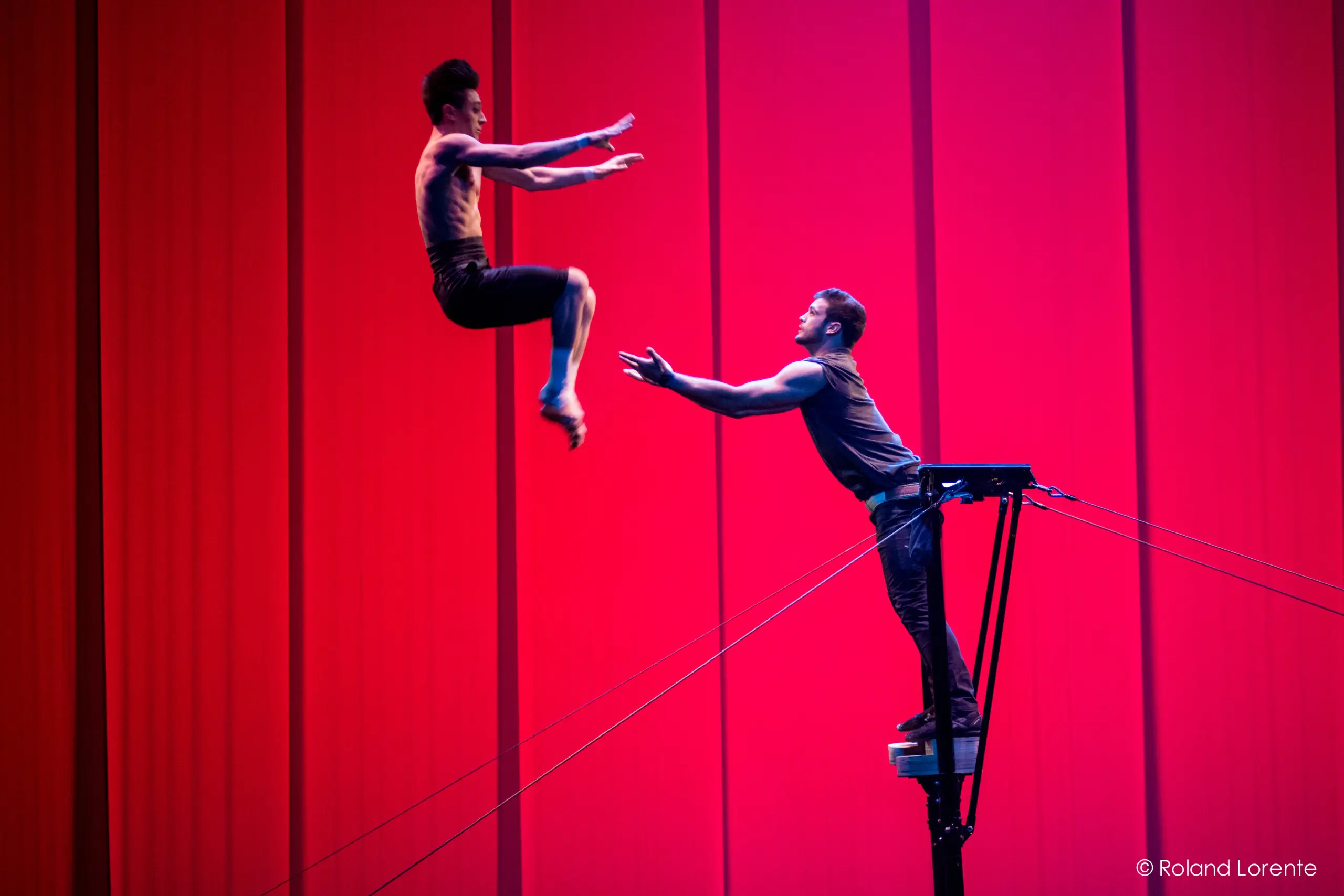 Deux acrobates, Louis Joyal et Samuel Renaud, réalisent une acrobatie : l'un saute dans les airs vers l'autre, qui se tient sur une plate-forme surélevée, les bras tendus, sur un fond rose-rouge vibrant.