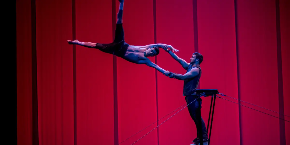 Deux acrobates masculins, Louis Joyal et Samuel Renaud, exécutent un numéro d'équilibre sur une plate-forme surélevée, sur un fond rouge vif. L'un des acrobates se penche spectaculairement vers l'extérieur, tendant la main à l'autre, qui le soutient en lui tenant le bras.