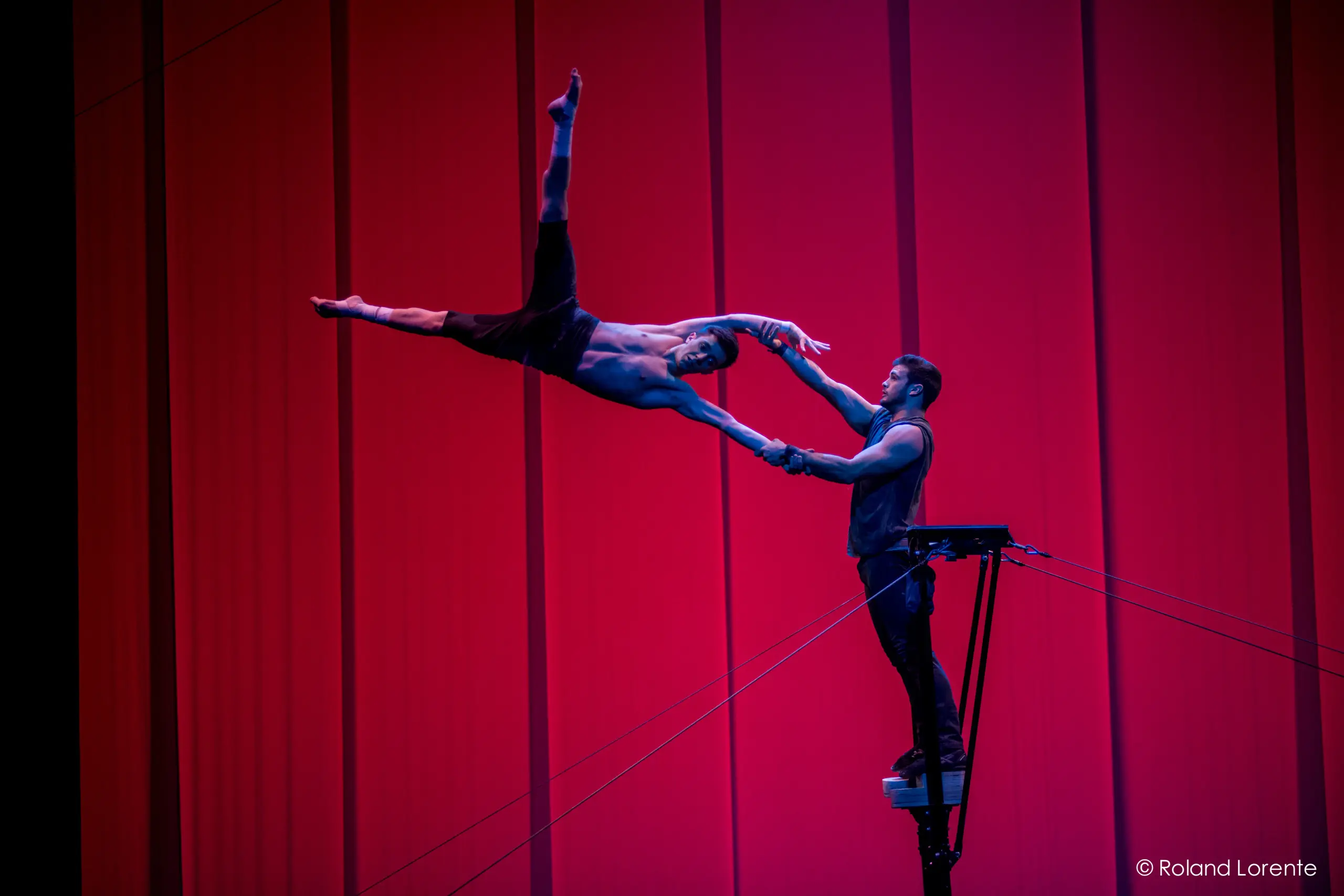 Deux acrobates masculins, Louis Joyal et Samuel Renaud, exécutent un numéro d'équilibre sur une plate-forme surélevée, sur un fond rouge vif. L'un des acrobates se penche spectaculairement vers l'extérieur, tendant la main à l'autre, qui le soutient en lui tenant le bras.