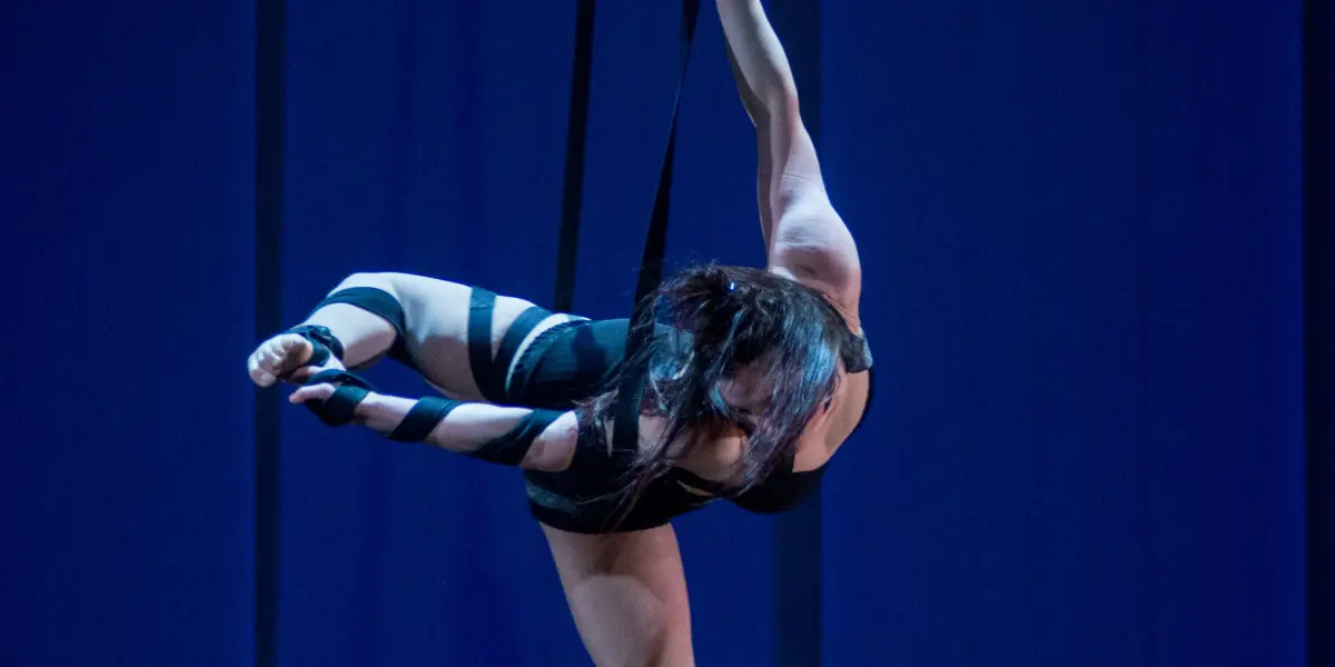 Une voltigeuse, Célia Milesi, en tenue noire, effectue une pose acrobatique en plein vol sur deux sangles suspendues, sur fond d'éclairage bleu, le corps gracieusement tendu.