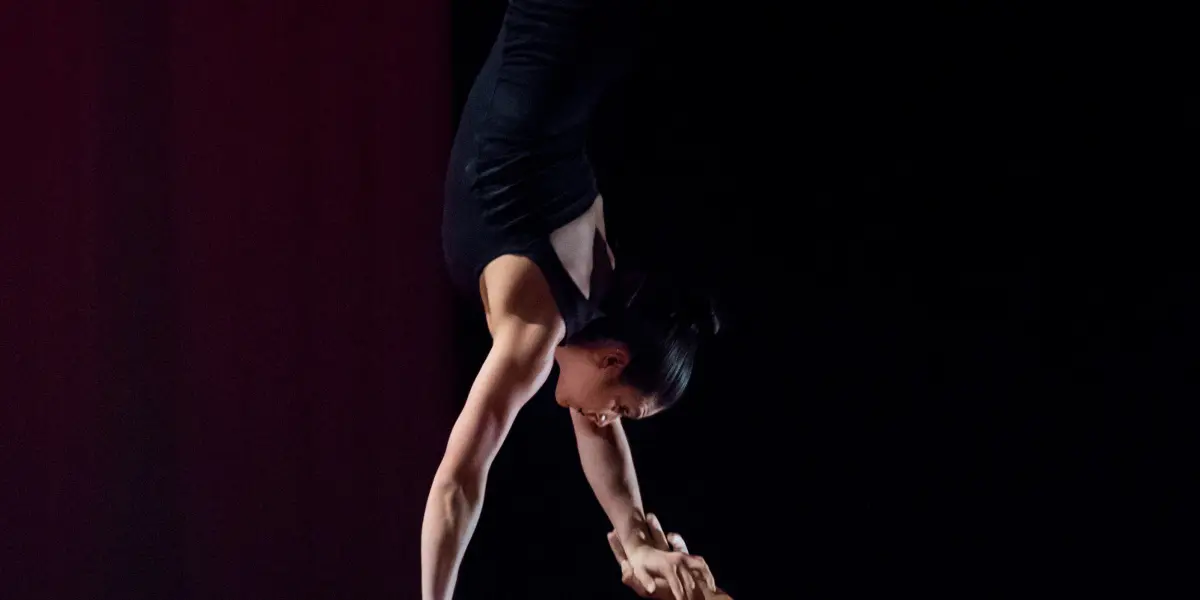 Un acrobate, Marlon Archer, tient la femme Jenay Espinosa en équilibre sur une main, la tête en bas. Elle prend une pose fendue, soutenue par son bras levé, tandis qu'il regarde vers le haut. L'arrière-plan sombre met en valeur leur athlétisme et leur équilibre.