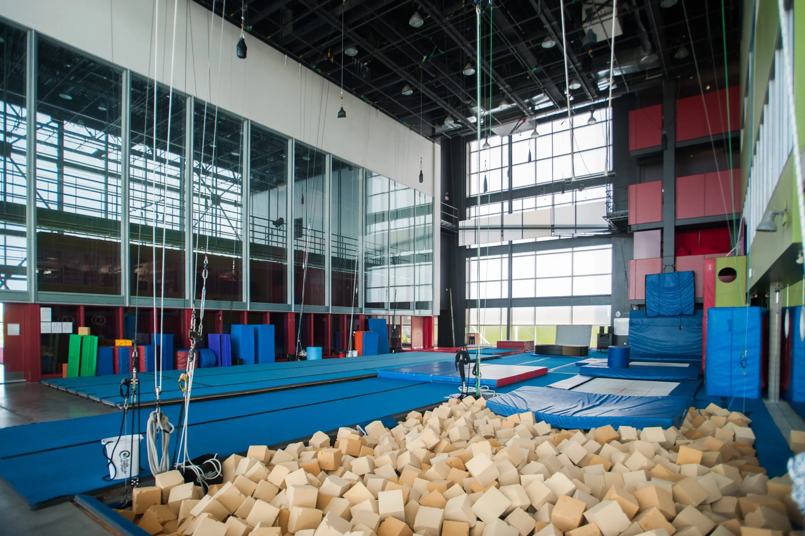 Un spacieux centre d'entraînement à la gymnastique en salle offre des services aux artistes de cirque professionnel. Il comprend une fosse à mousse, des tapis bleus, des cordes à grimper, des poutres d'équilibre et de grandes fenêtres. Les murs sont recouverts d'un rembourrage coloré pour plus de sécurité.