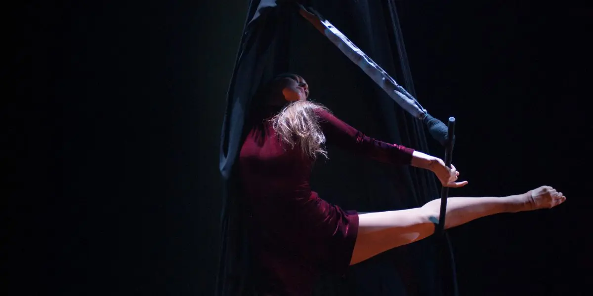 L'artiste aérienne Catherine Beaudet, vêtue d'une tenue bordeaux, se suspend gracieusement et prend la pose sur un tissu de soie noire, une jambe tendue à l'horizontale, sur une scène sombre sous un éclairage dramatique.