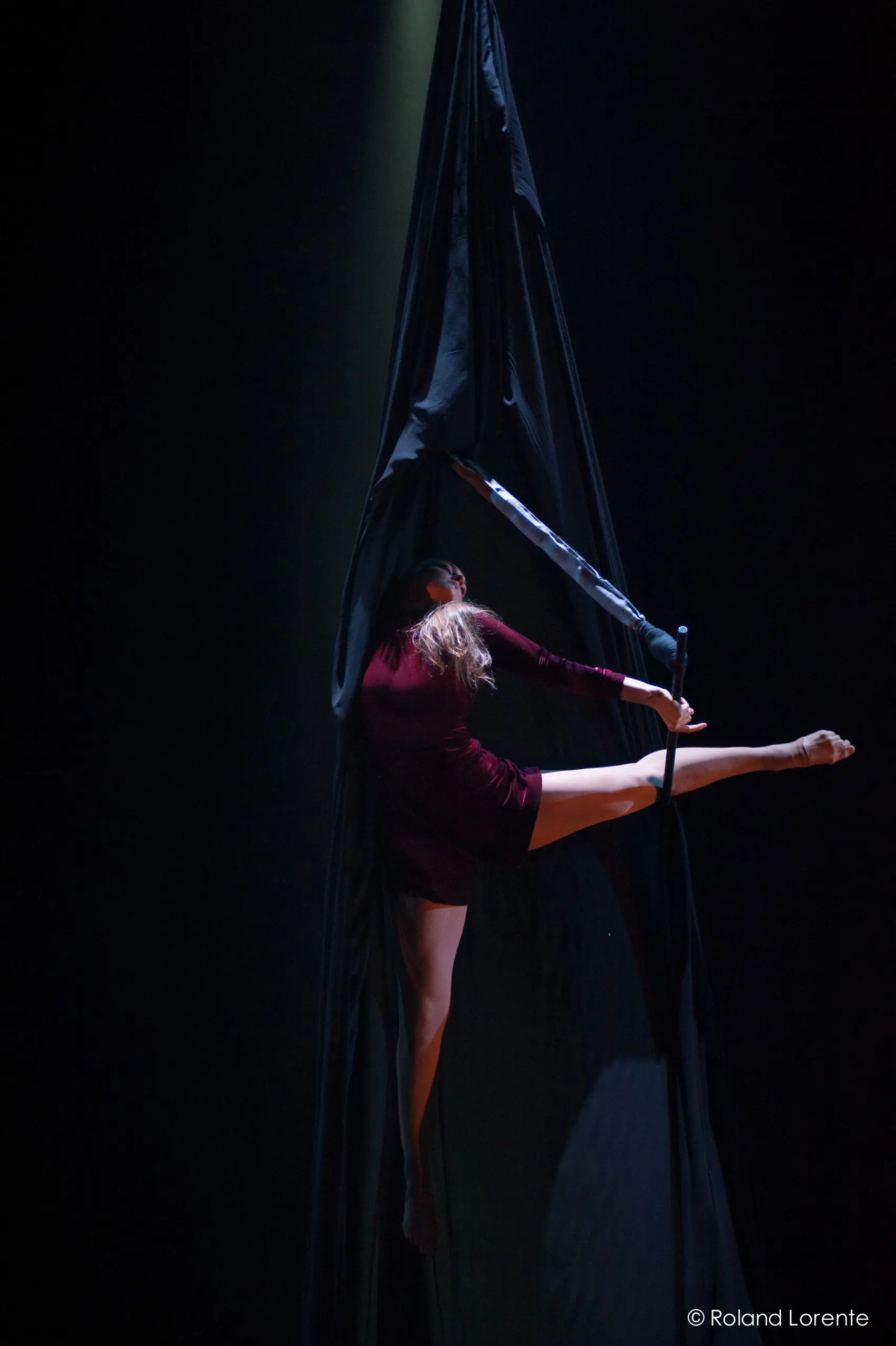 L'artiste aérienne Catherine Beaudet, vêtue d'une tenue bordeaux, se suspend gracieusement et prend la pose sur un tissu de soie noire, une jambe tendue à l'horizontale, sur une scène sombre sous un éclairage dramatique.