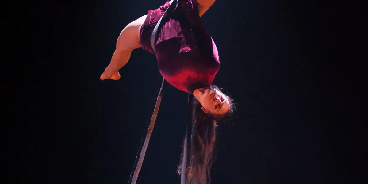 Catherine Beaudet, une artiste aérienne vêtue d'un costume bordeaux, exécute une pose acrobatique à l'envers sur un trapèze suspendu, sous les feux de la rampe sur un fond sombre.