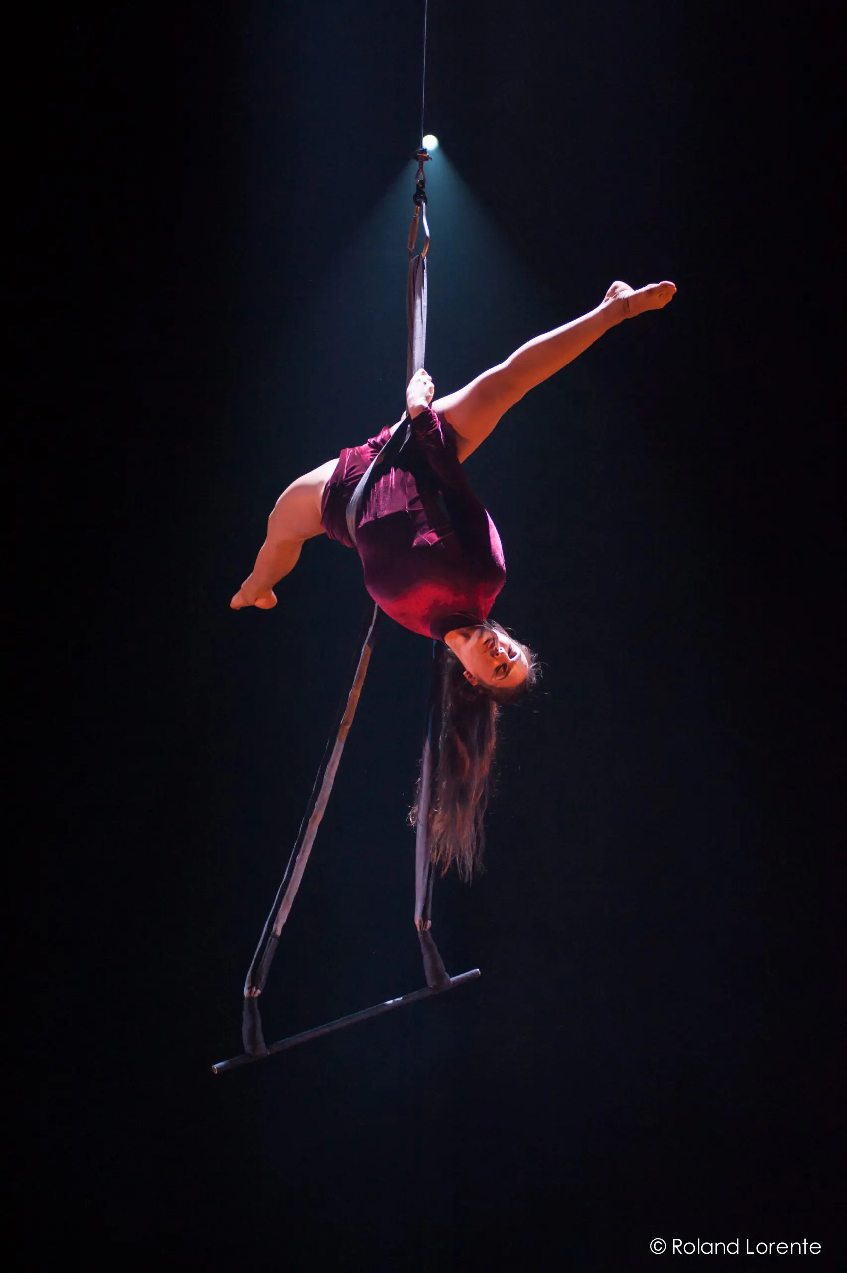 Catherine Beaudet, une artiste aérienne vêtue d'un costume bordeaux, exécute une pose acrobatique à l'envers sur un trapèze suspendu, sous les feux de la rampe sur un fond sombre.