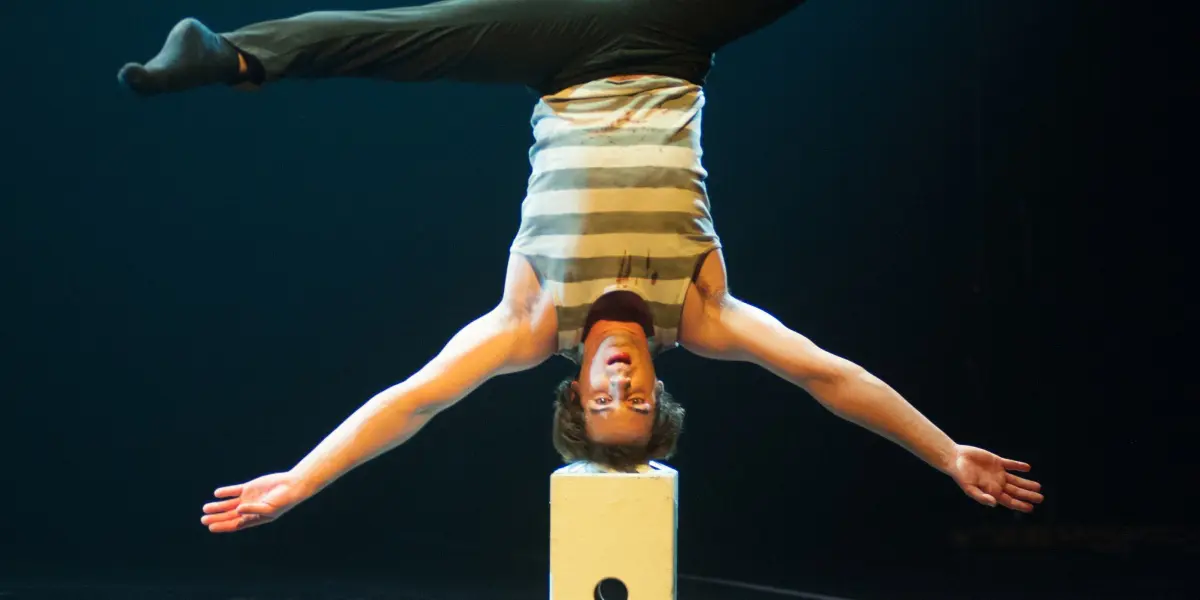 Florian Jeannot se tient en équilibre sur un bloc blanc, les jambes tendues en grand écart, sur une scène sombre et réfléchissante.