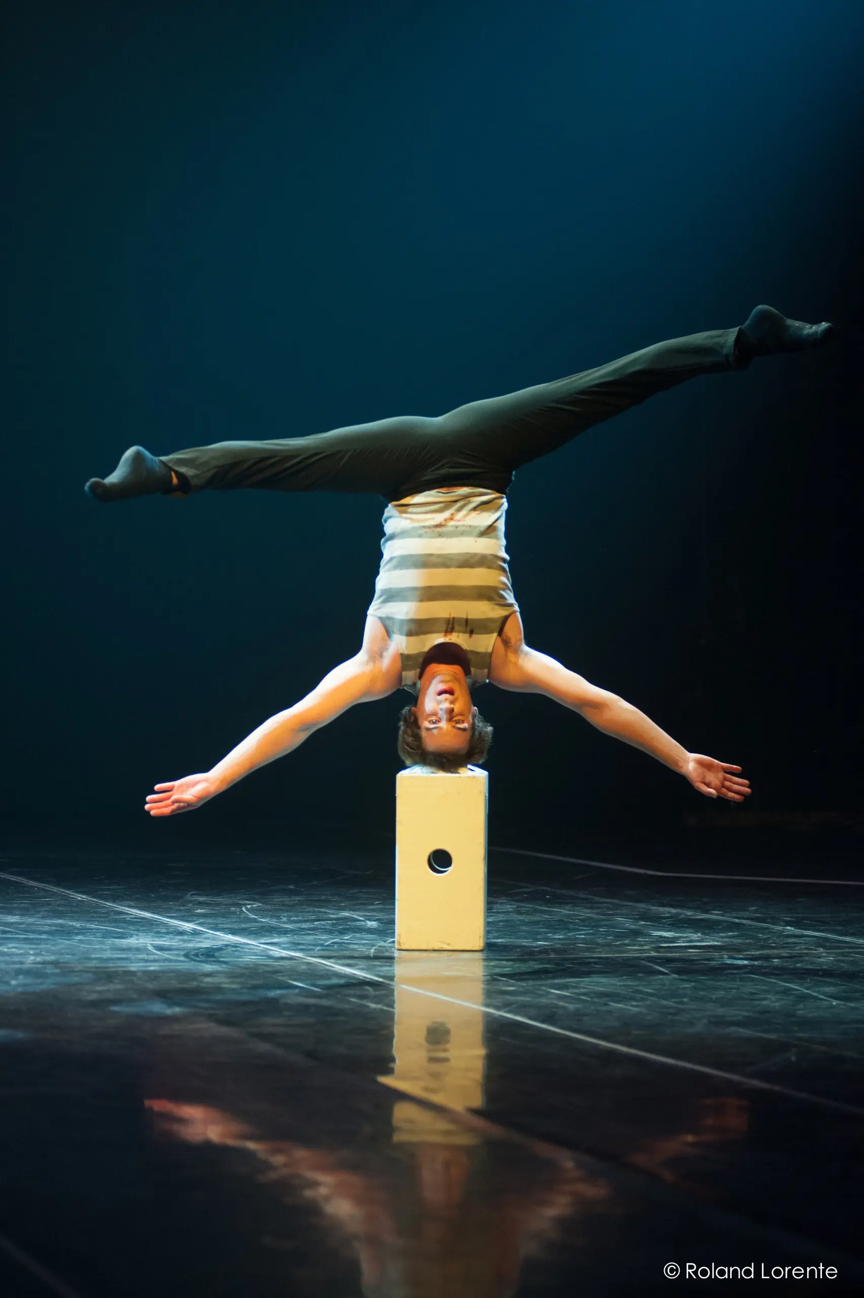 Florian Jeannot se tient en équilibre sur un bloc blanc, les jambes tendues en grand écart, sur une scène sombre et réfléchissante.