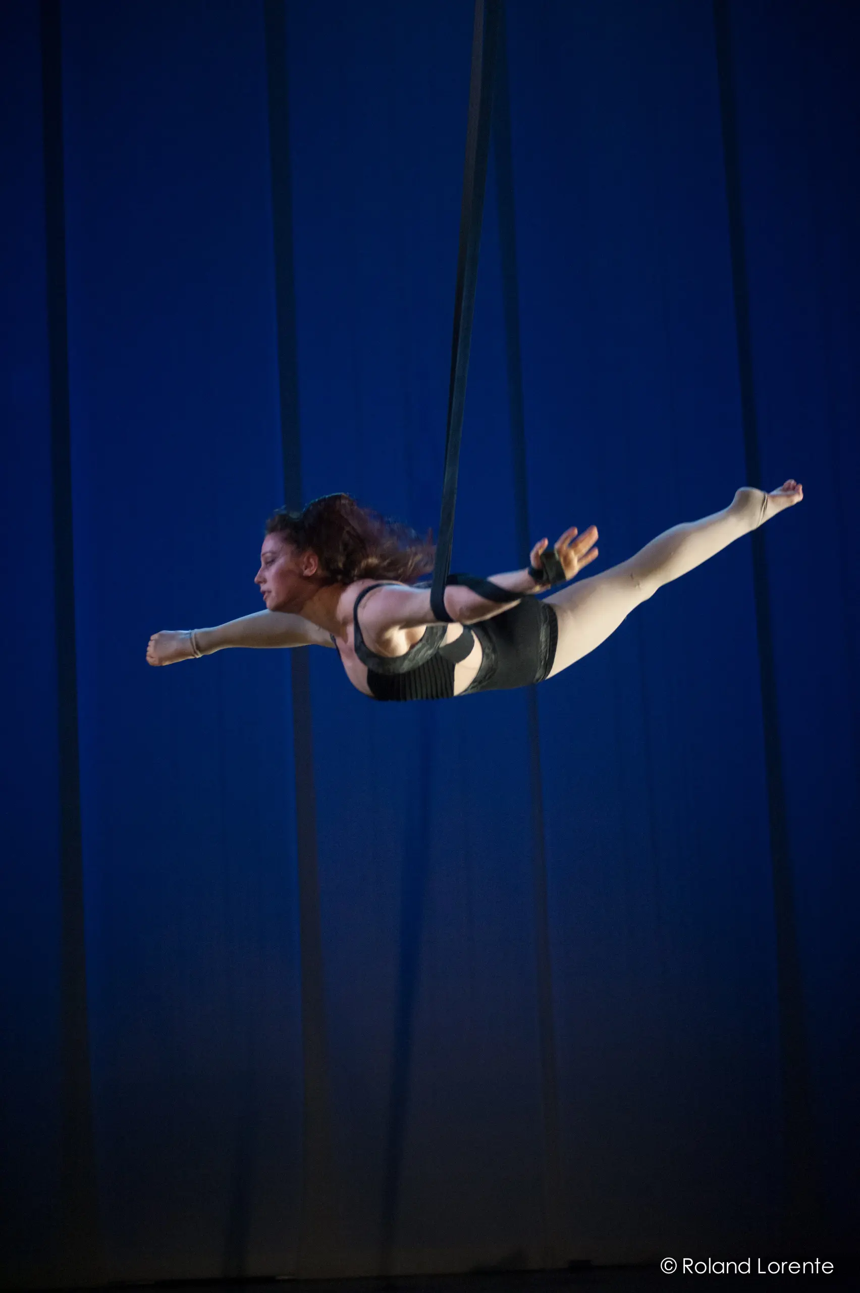 Une voltigeuse, Célia Milesi, en tenue noire, effectue une pose horizontale suspendue à des sangles aériennes sur un fond bleu, semblant voler avec un bras tendu vers l'avant.