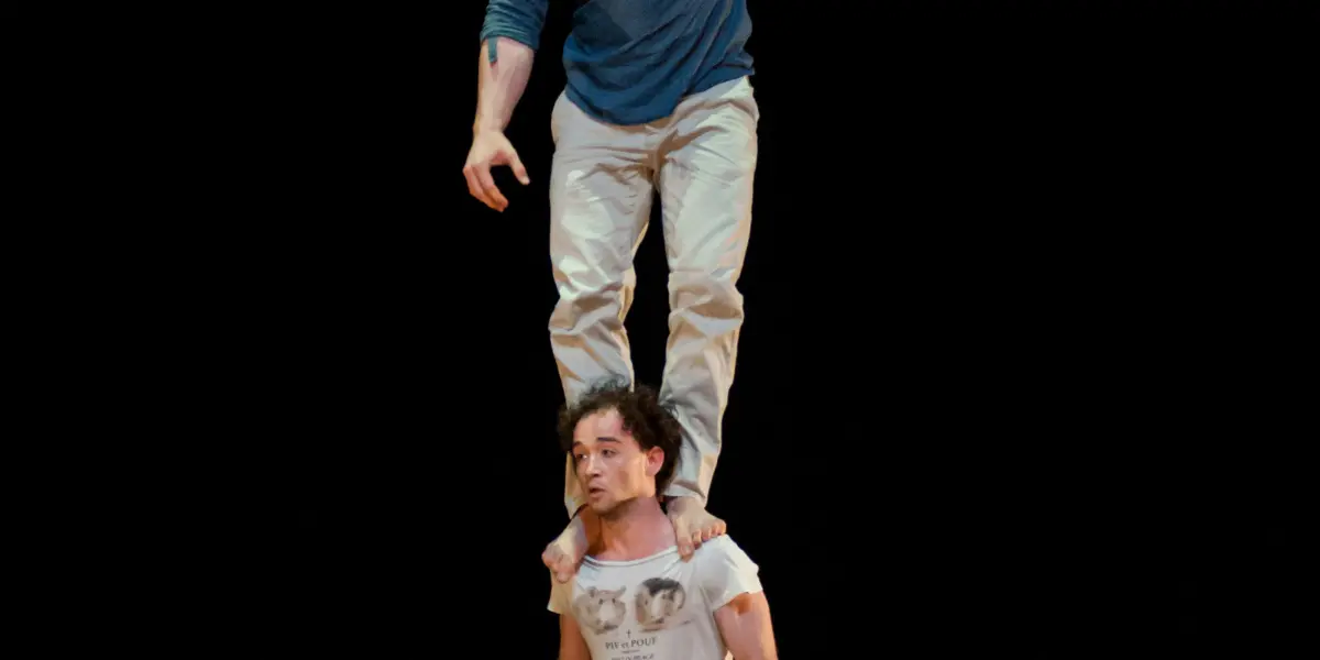 Deux artistes masculins, Julius Bitterling et César Mispelon, sont en équilibre sur scène ; l'un se tient pieds nus sur les épaules de l'autre. Tous deux portent des vêtements décontractés sur un fond noir.