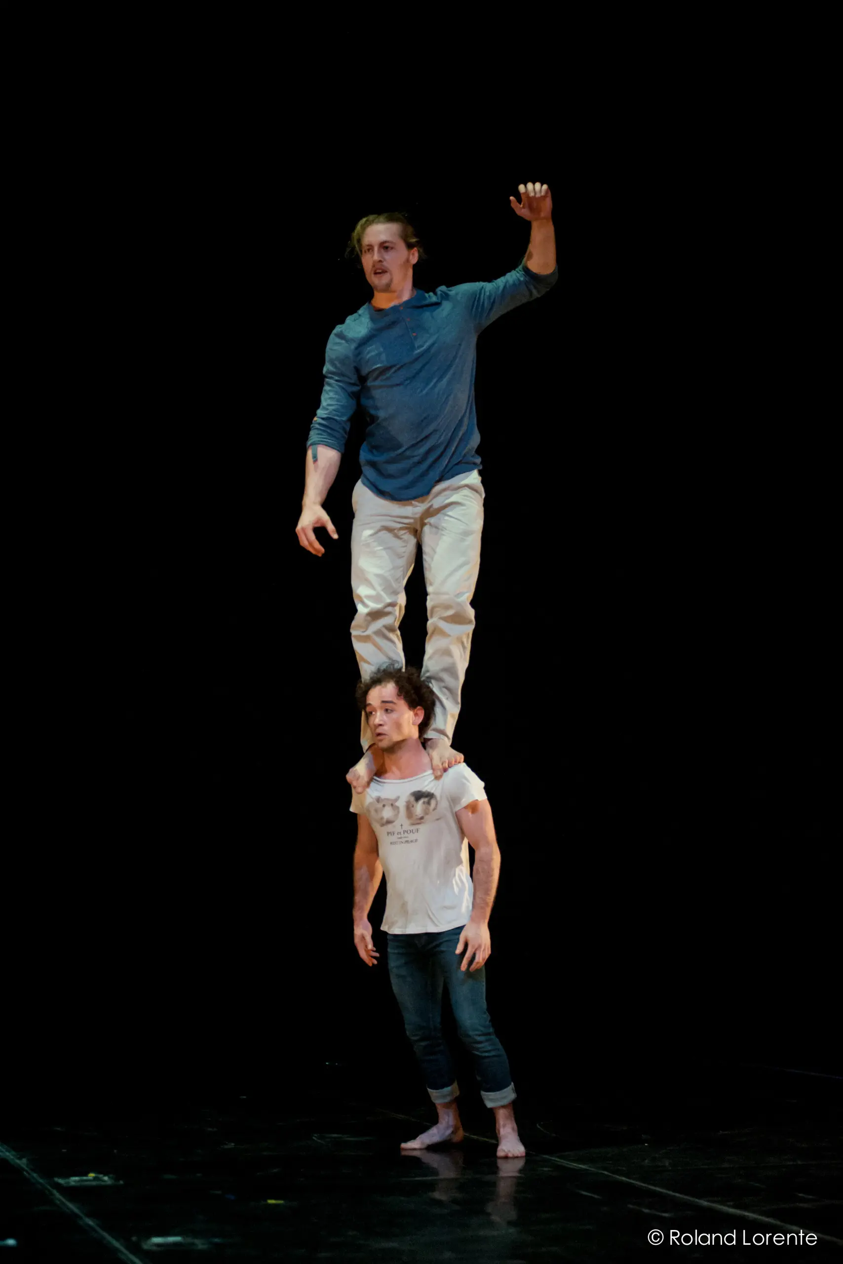 Deux artistes masculins, Julius Bitterling et César Mispelon, sont en équilibre sur scène ; l'un se tient pieds nus sur les épaules de l'autre. Tous deux portent des vêtements décontractés sur un fond noir.