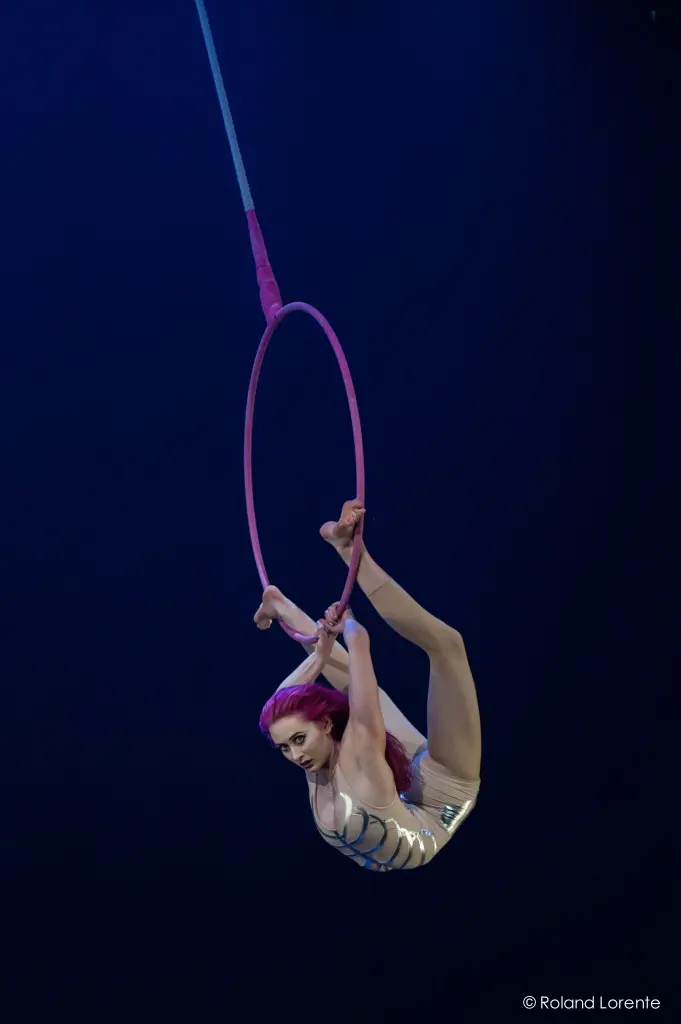 L'artiste aérienne Angela Mcllroy-Wagar, aux cheveux roses et vêtue d'un costume blanc, se suspend la tête en bas à un cerceau rose, en arquant le dos et en tenant le cerceau avec ses pieds sur une scène éclairée en bleu foncé.