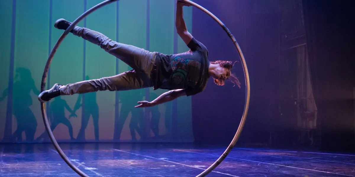 Cory Marsh se tient en équilibre latéral à l'intérieur d'un grand cerceau métallique sur une scène, avec un éclairage dramatique et des ombres projetées sur la toile de fond.