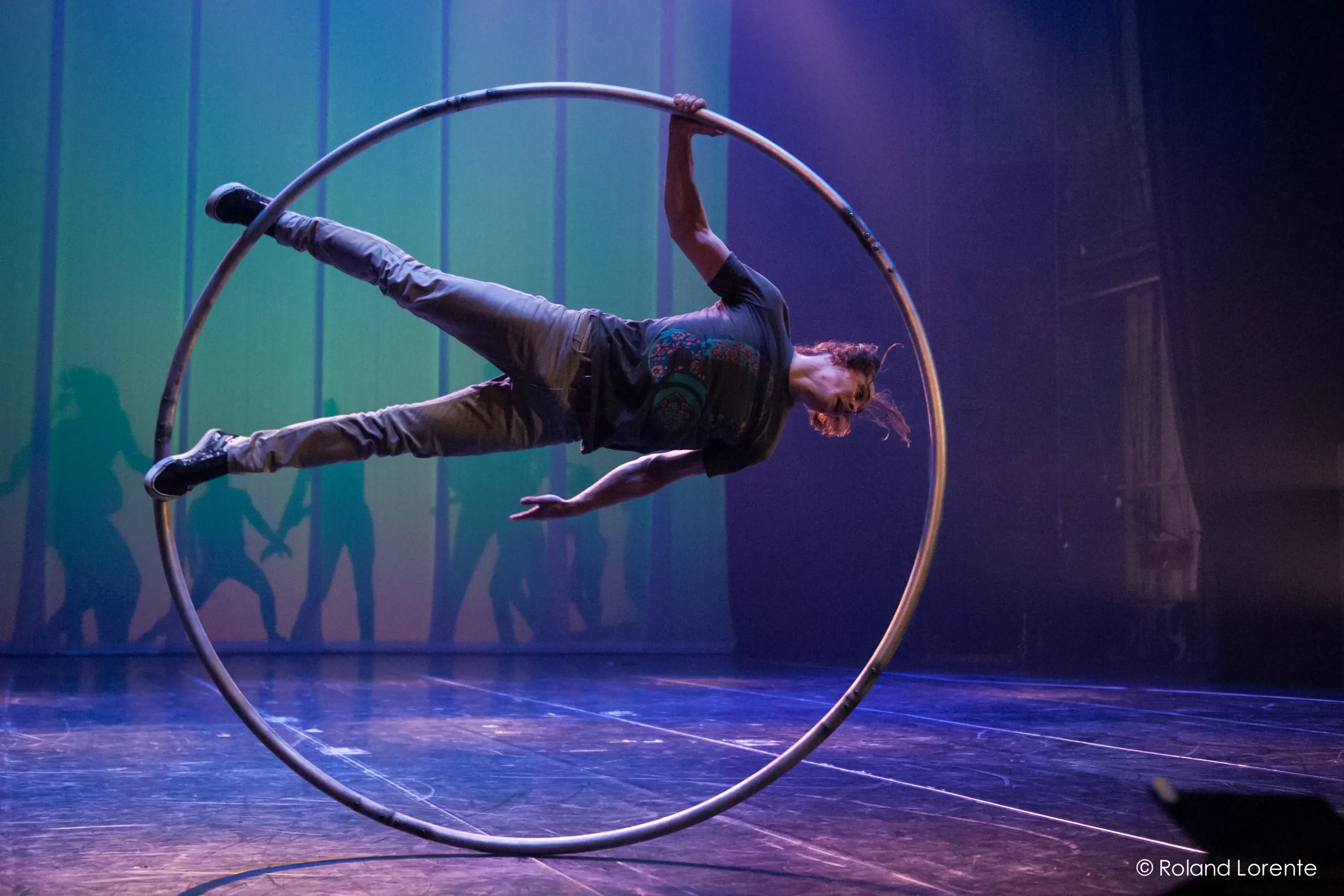 Cory Marsh se tient en équilibre latéral à l'intérieur d'un grand cerceau métallique sur une scène, avec un éclairage dramatique et des ombres projetées sur la toile de fond.