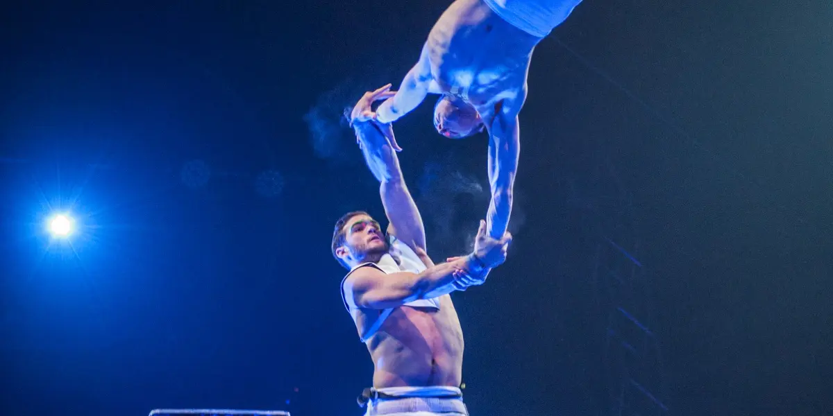 Deux acrobates, Louis Joyal et Samuel Renaud, en tenue blanche, exécutent un numéro de trapèze de haut vol sous les lumières bleues de la scène ; l'un se tient sur une plate-forme tandis que l'autre est dans les airs, saisissant les mains de son partenaire en plein vol.