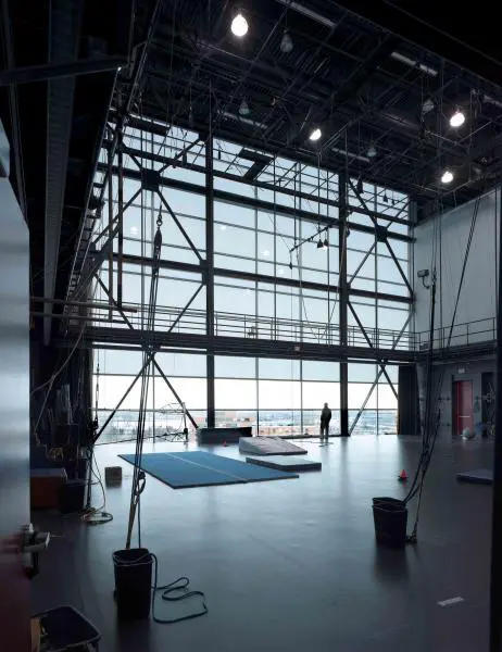 Un centre d'entraînement intérieur spacieux avec de hauts plafonds, de grandes fenêtres, des tapis de sol, des cordes suspendues et une personne près des fenêtres - parfait pour les services aux artistes de cirque professionnels à la recherche d'un espace d'entraînement inspirant.