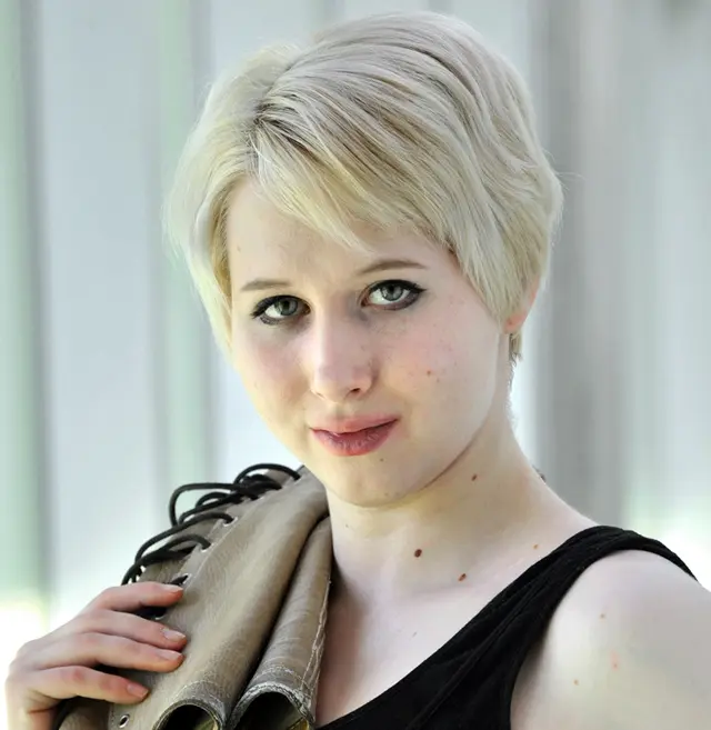 Une jeune personne aux cheveux courts et blonds et à la peau claire tient une veste beige pliée sur son épaule, porte un haut noir sans manches et regarde directement l'appareil photo avec un léger sourire. L'arrière-plan légèrement flou met en valeur la pose détendue d'Alma Buholzer.