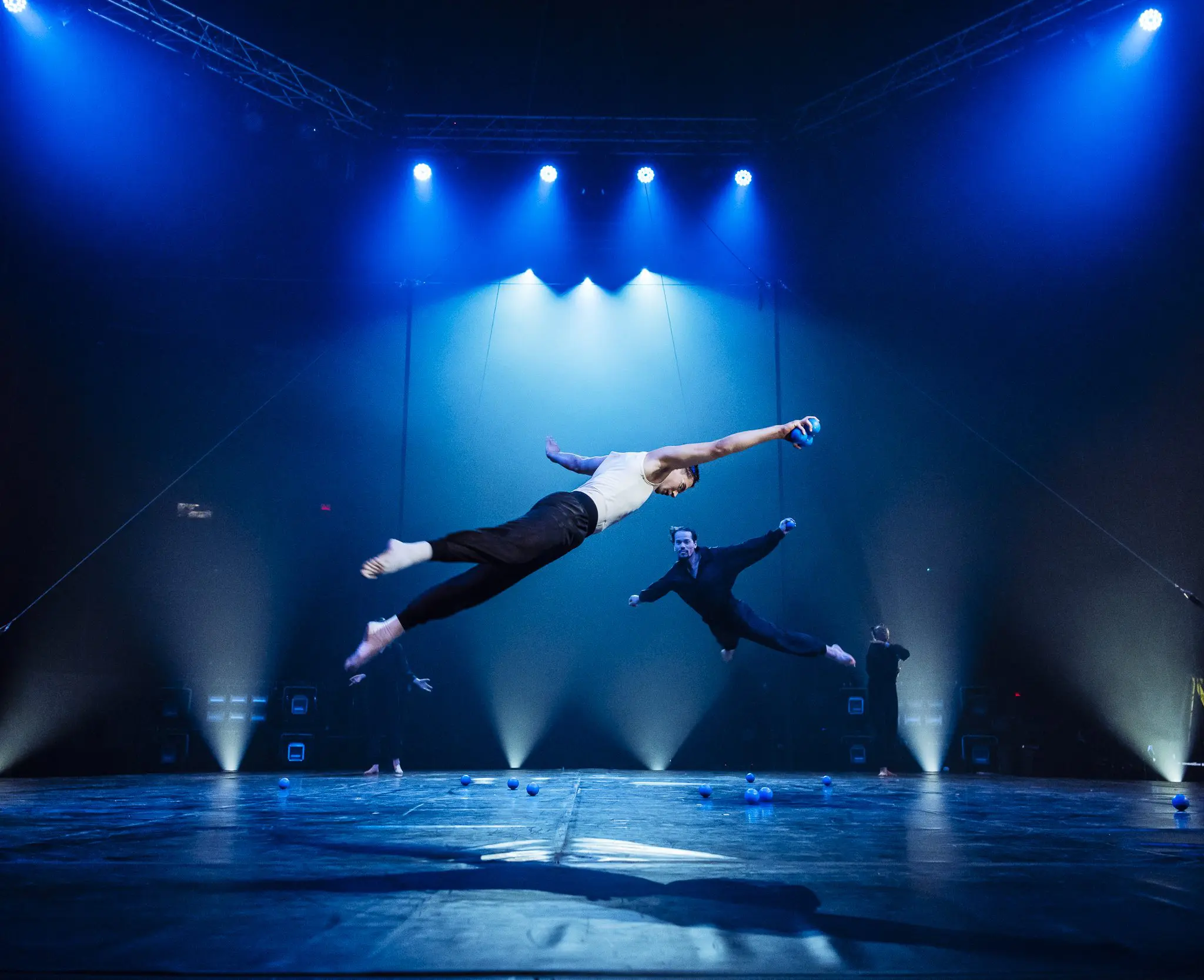 Deux acrobates, tous deux diplomés de l'ECN avant 2007, bondissent sur une scène sous des projecteurs bleus, bras et jambes tendus, tandis que d'autres artistes et équipements scéniques sont visibles à l'arrière-plan.