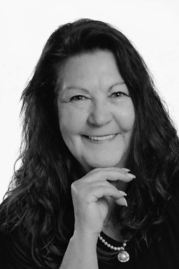 Marie-Reine Forget, aux longs cheveux ondulés, sourit chaleureusement à l'appareil photo. Elle porte un haut sombre et un collier de perles, sa main se posant délicatement sur son menton. La photo en noir et blanc présente un fond clair.