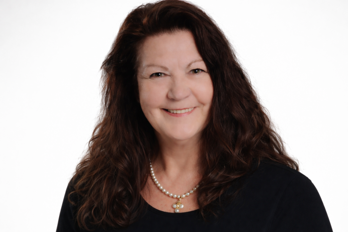 Marie-Reine Forget, une femme aux longs cheveux bruns, portant un haut noir et un collier de perles, sourit à l'appareil photo sur un fond blanc uni.