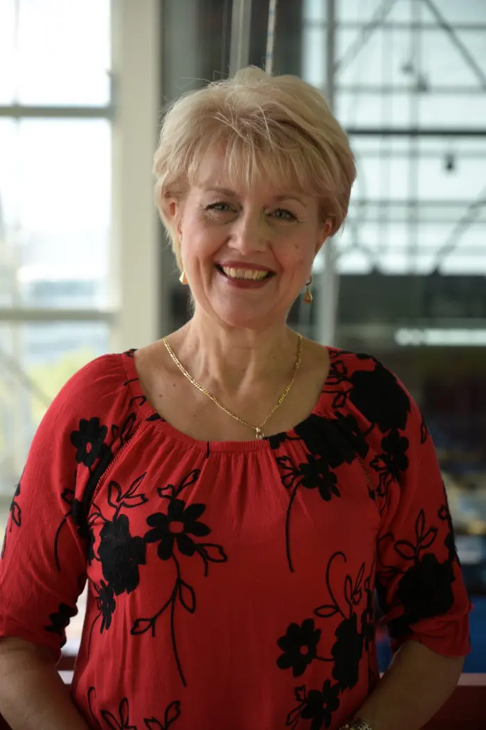 Une femme souriante aux cheveux blonds courts, ressemblant à Eugenia Nicolae, porte un chemisier rouge avec des motifs floraux noirs, un collier en or et des boucles d'oreilles. Elle se tient à l'intérieur, près de grandes fenêtres, avec un arrière-plan légèrement flou.