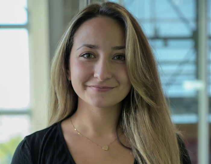 Shany Bergeron-Roy, une femme aux longs cheveux châtain clair, sourit doucement à la caméra. Elle porte un haut noir et un pendentif doré en forme de cœur. L'arrière-plan est composé de grandes fenêtres et d'un décor intérieur flou.